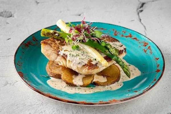 A beautifully plated dish of grilled fish served with asparagus, microgreens, and creamy sauce over sliced potatoes. The dish is presented on a vibrant turquoise plate.