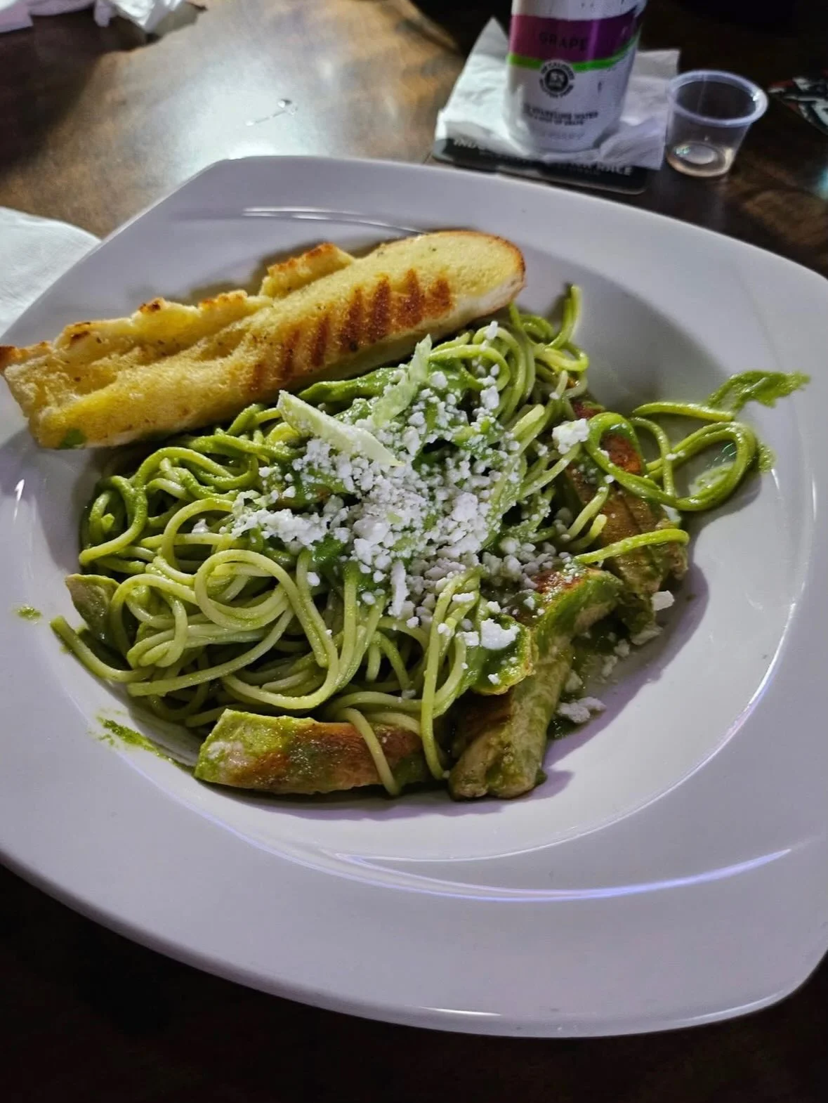 A plate of green pesto spaghetti topped with cheese and served with garlic bread on the side.