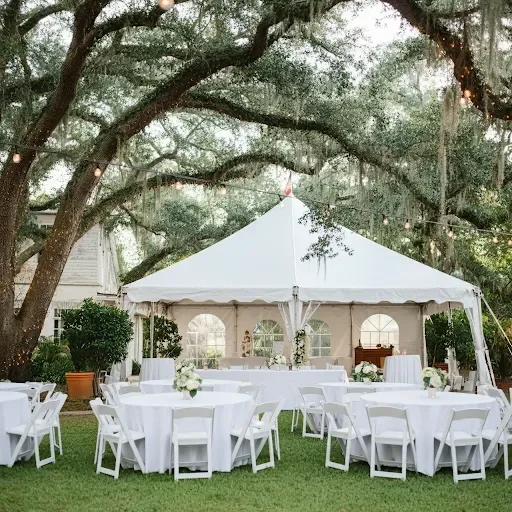 An outdoor event space with white tables arranged under a large tent, surrounded by trees adorned with lights. The setting is elegant and inviting.