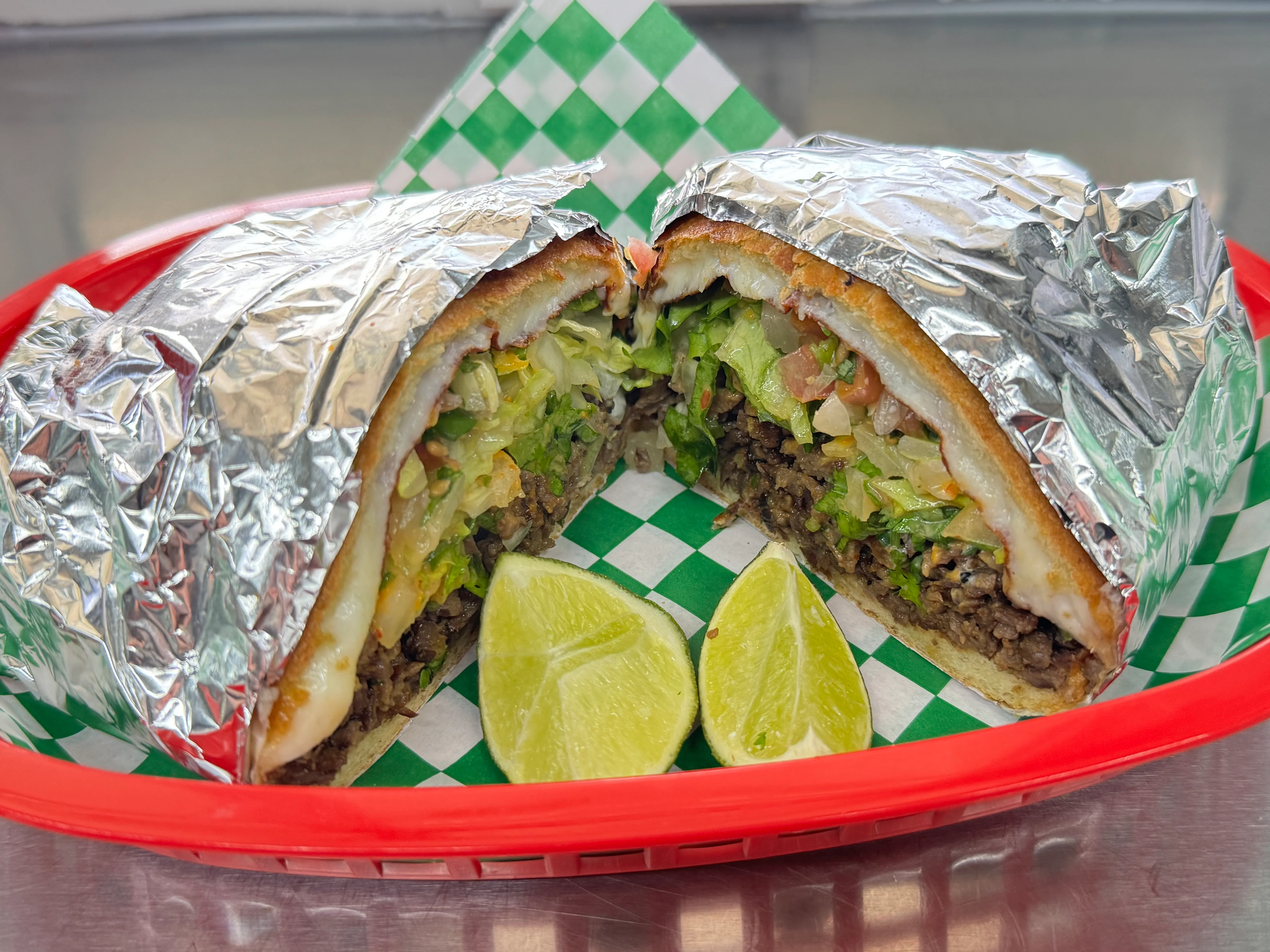 A halved, stuffed tortilla wrapped in foil, showcasing layers of meat, lettuce, and tomatoes, accompanied by lime wedges in a red basket.