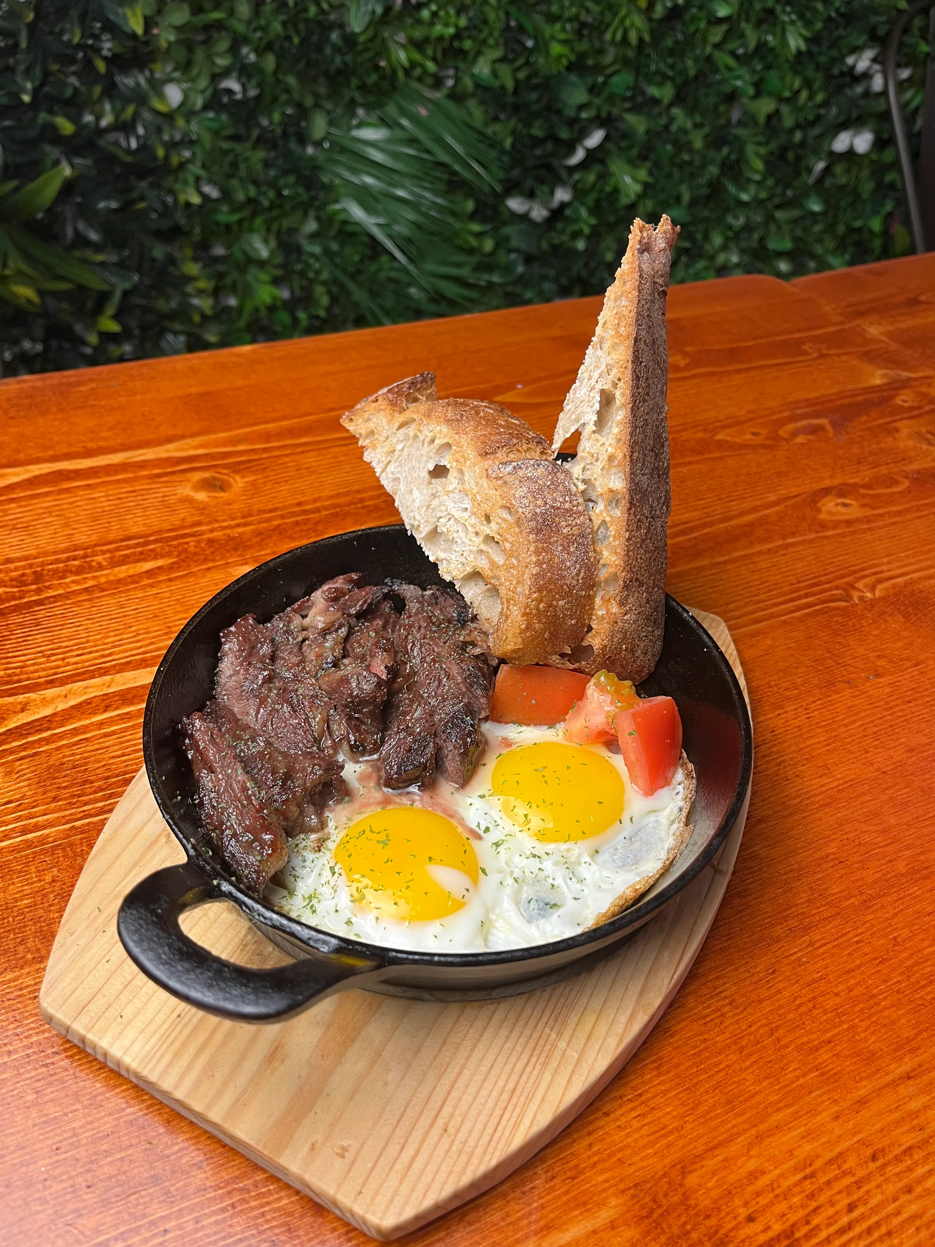 A skillet meal featuring two sunny-side-up eggs, strips of beef, chopped tomatoes, and toasted bread, served on a wooden board.