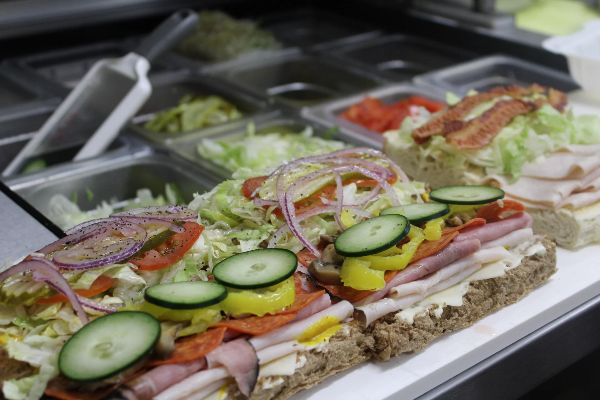 A long sandwich filled with layers of deli meats, vegetables, and toppings on a bread base, displayed on a countertop.