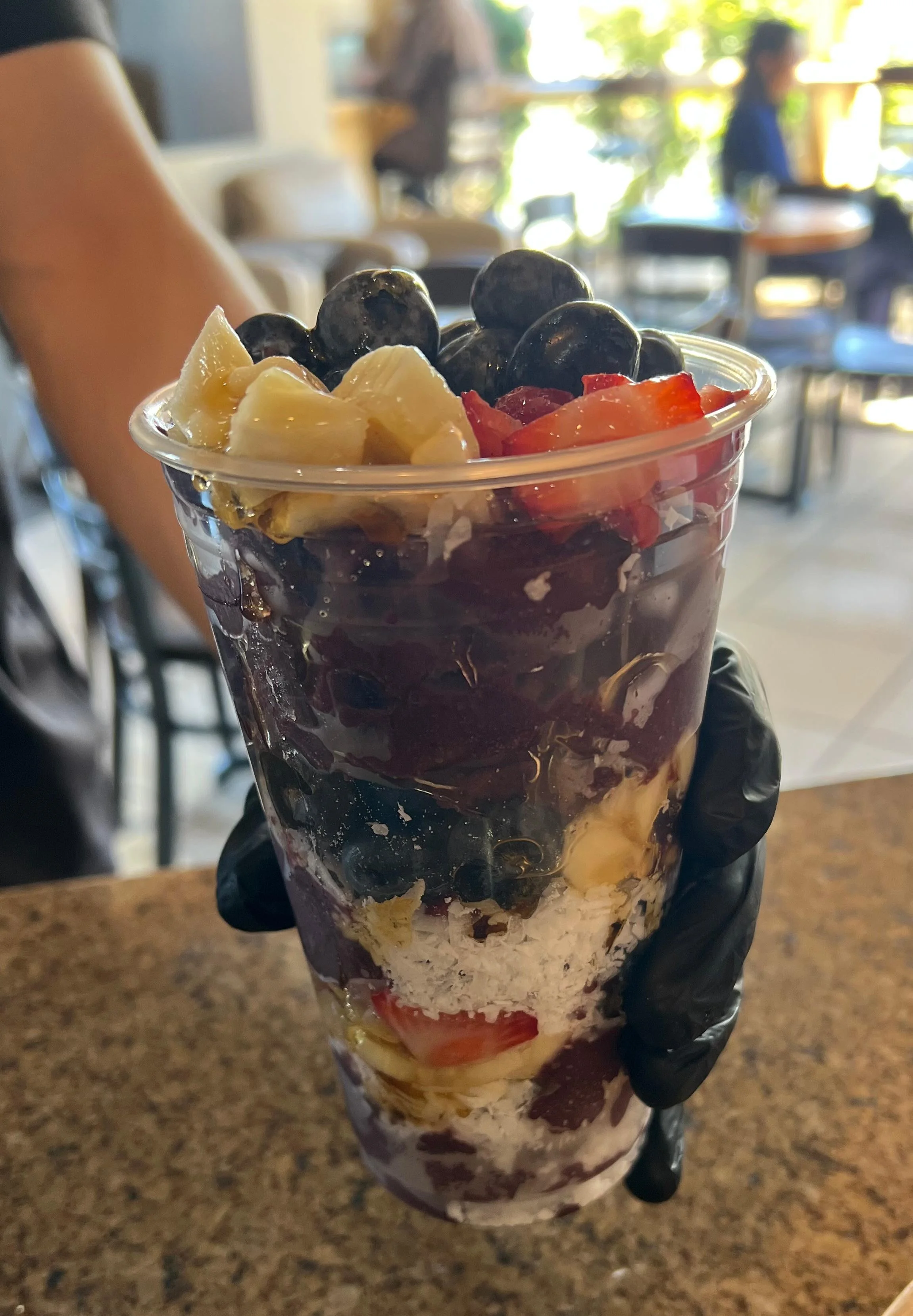 A tall cup filled with layers of fruits including blueberries, bananas, and strawberries, along with a creamy layer, held by a hand in a black glove.