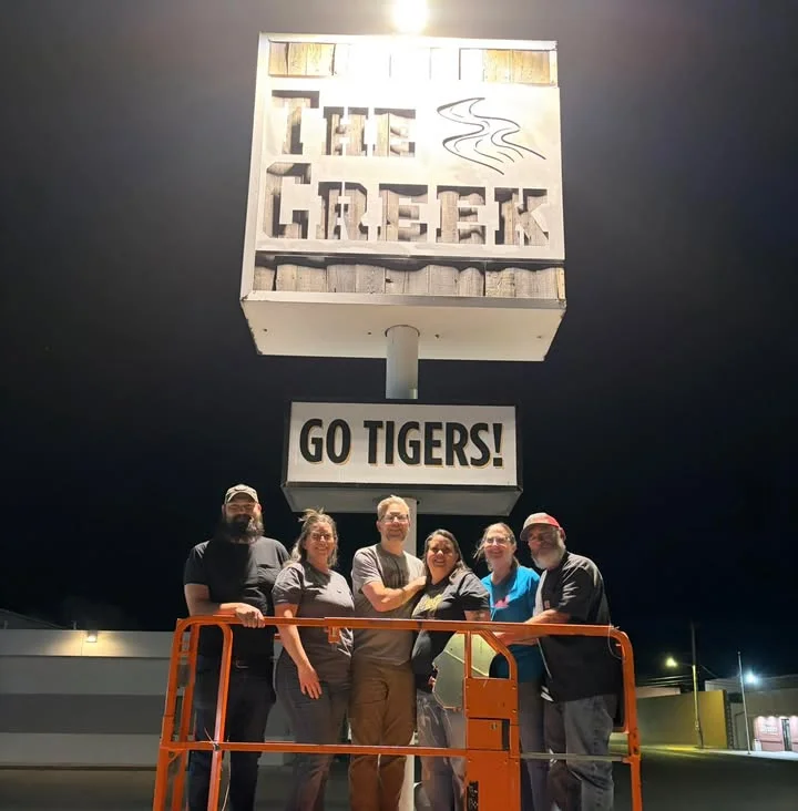 A group of six people standing in front of a restaurant sign that reads 'The Creek' and 'Go Tigers!' at night.
