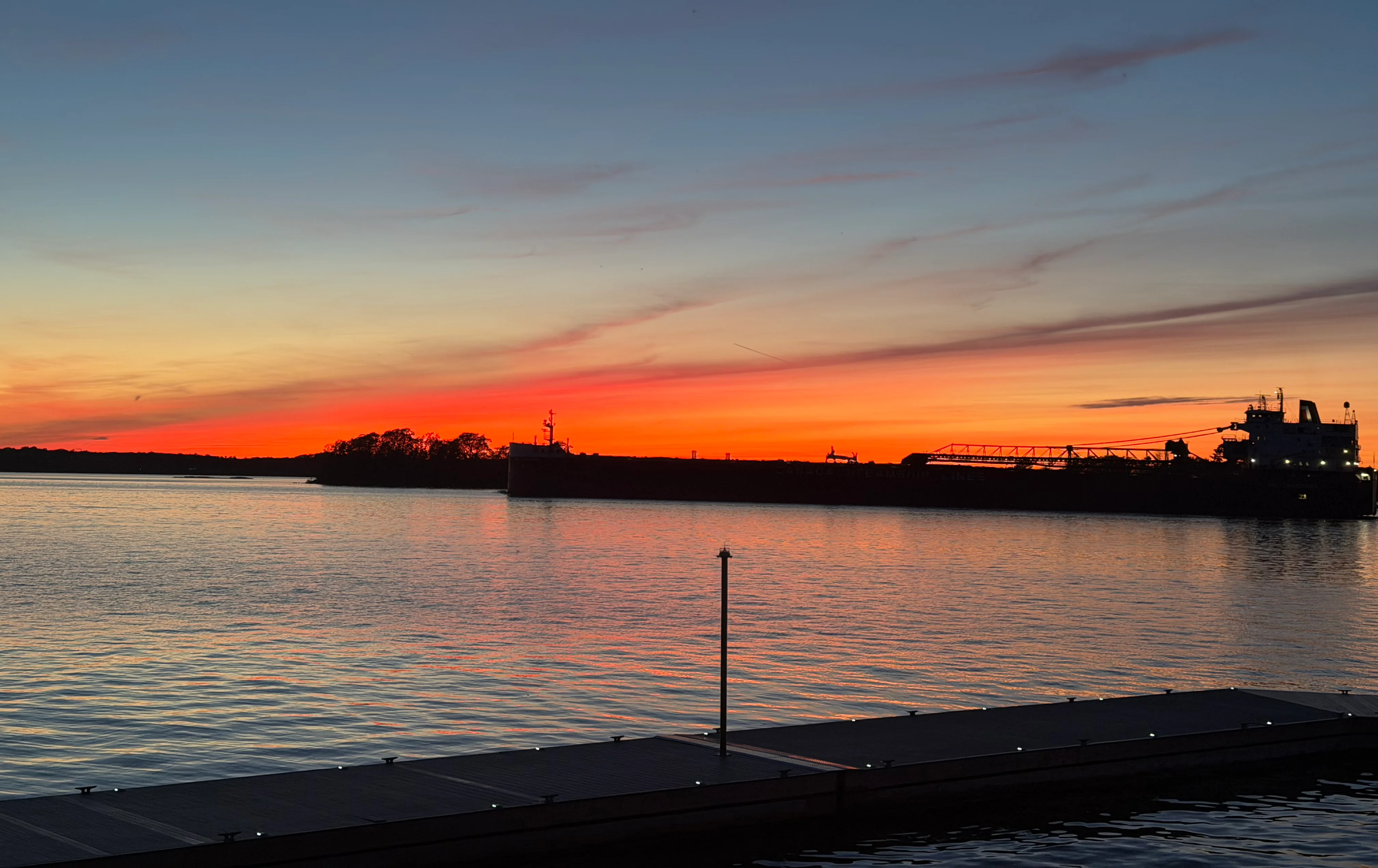Vibrant sunset over calm water with silhouettes of trees and a ship