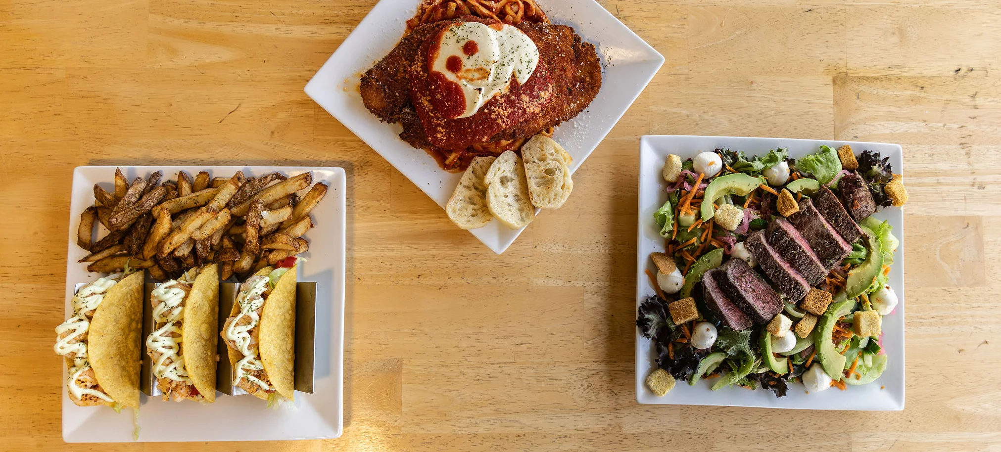 Plates of tacos, chicken parmesan, and steak salad on a wooden table