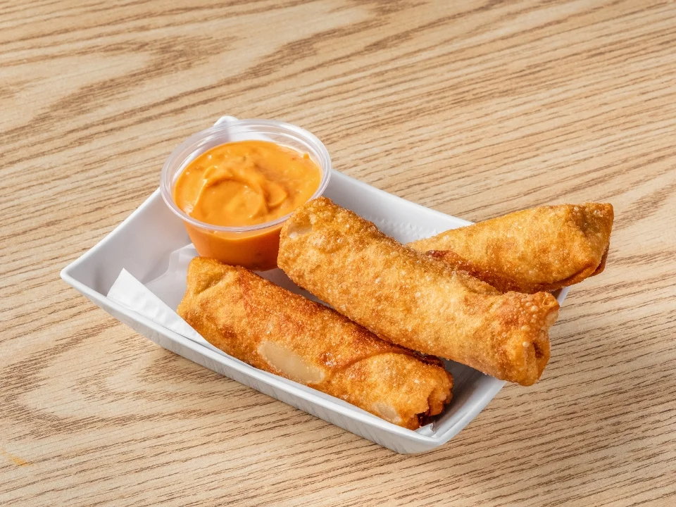 A plate with three crispy eggrolls served alongside a cup of dipping sauce, placed on a wooden table.