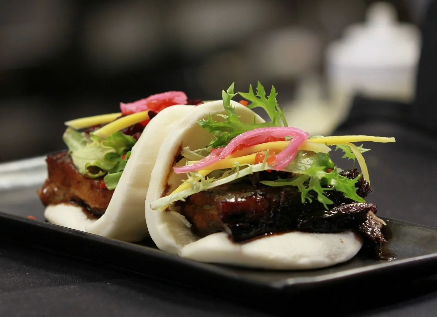 Two soft, white bao buns filled with thick slices of glazed pork belly, fresh greens, julienned yellow vegetables, and pickled red onions, served on a narrow black plate.