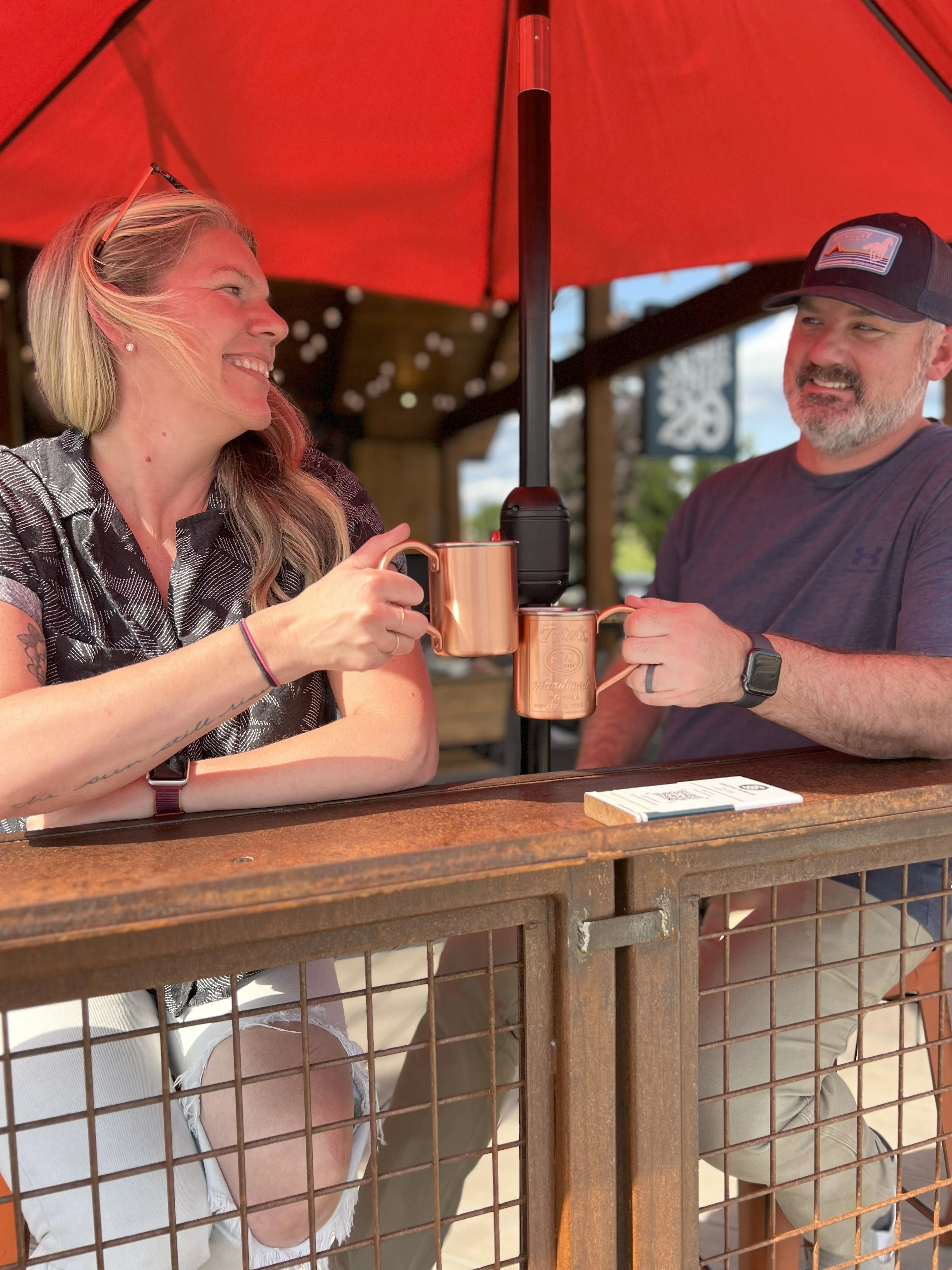 Two friends toasting with mugs under a red umbrella at a restaurant patio