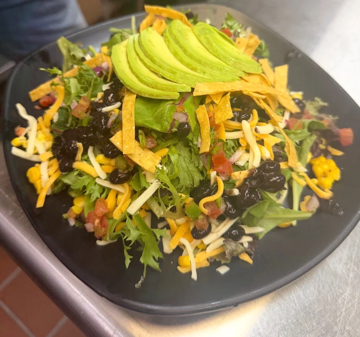A colorful salad served on a black plate, topped with tortilla strips and avocado slices.