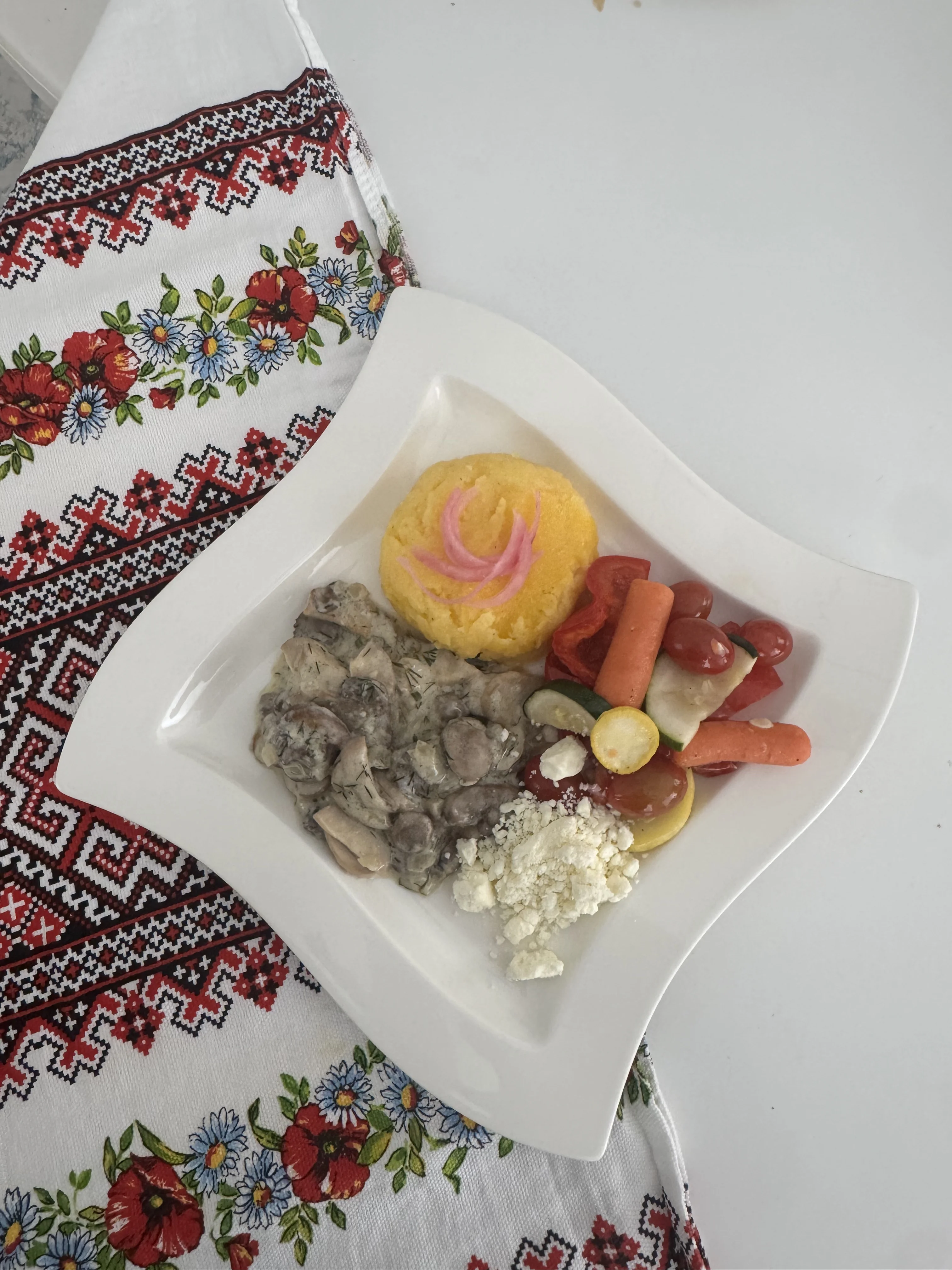 A plated meal with a serving of creamy chicken, polenta, assorted vegetables, and fresh cheese, presented on a decorative table setting.