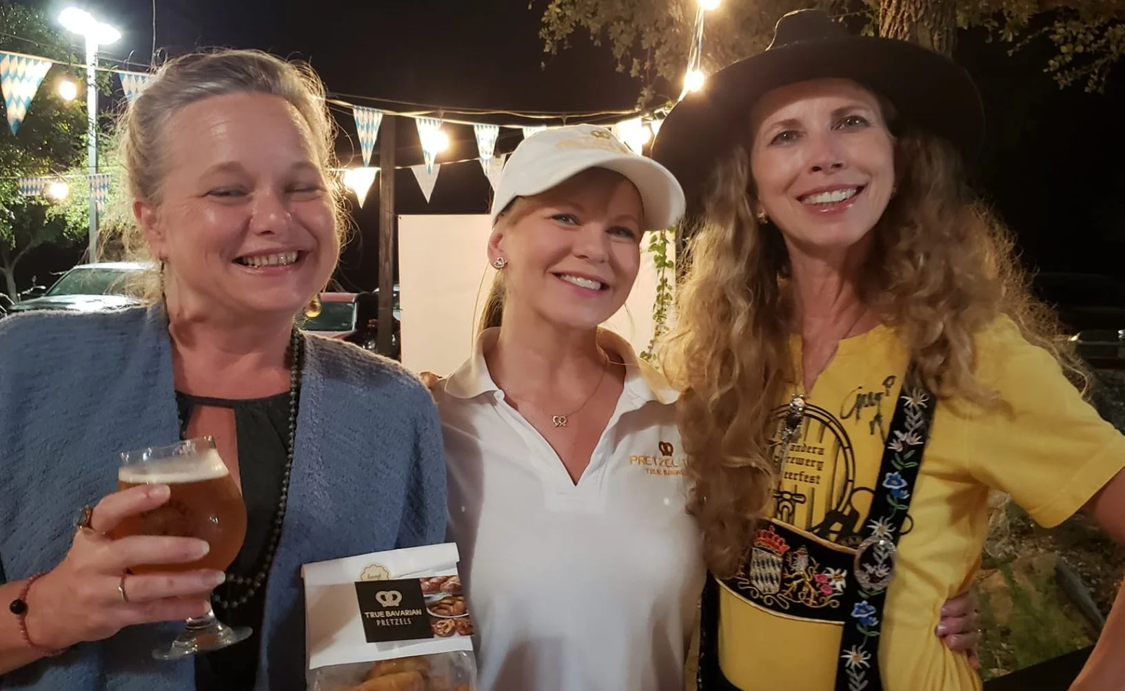 Three women smiling at a festive outdoor event with pretzels and drinks.