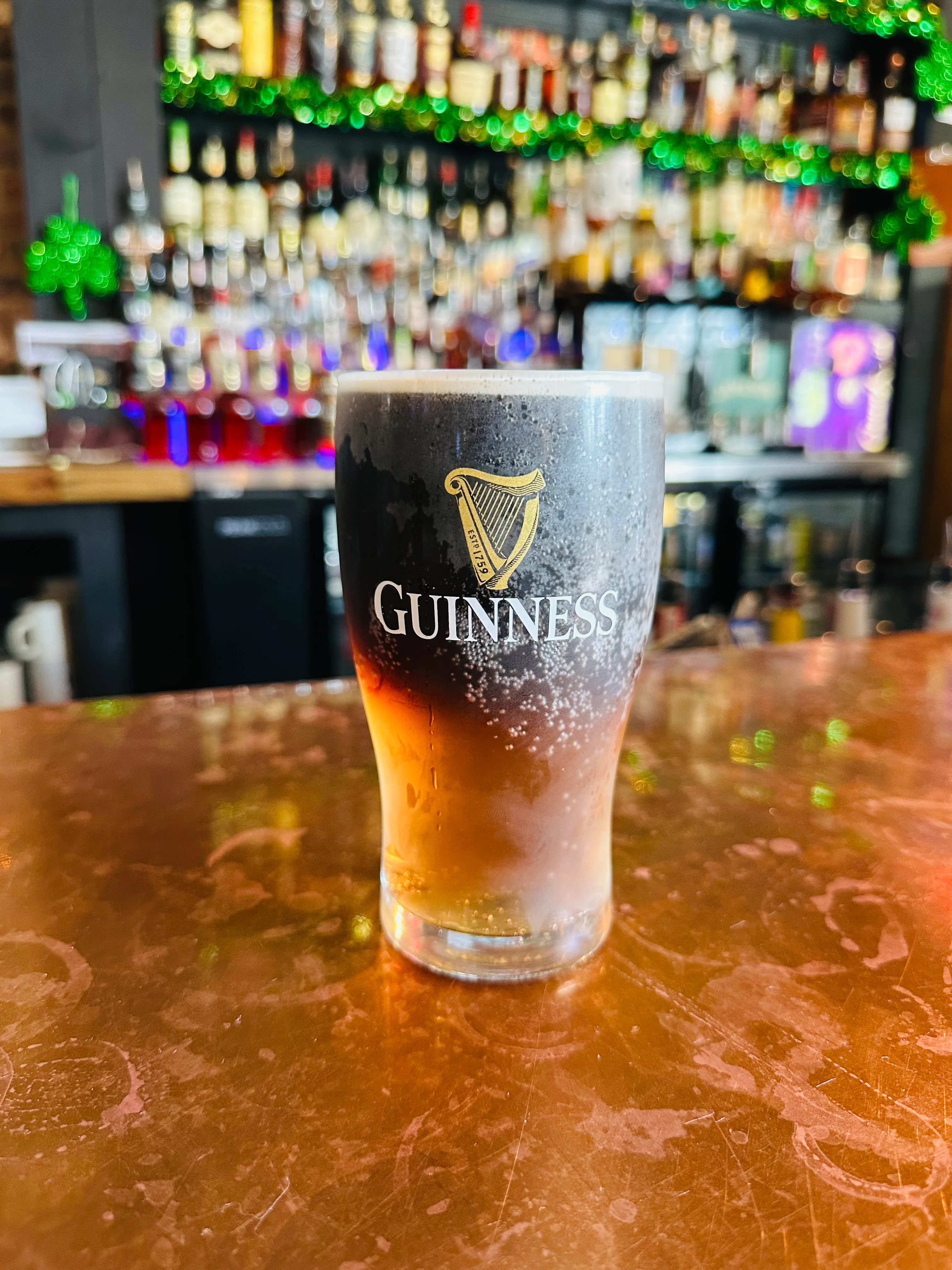 A pint of Guinness beer with a frothy head placed on a copper bar top, with a blurry backdrop of liquor bottles.