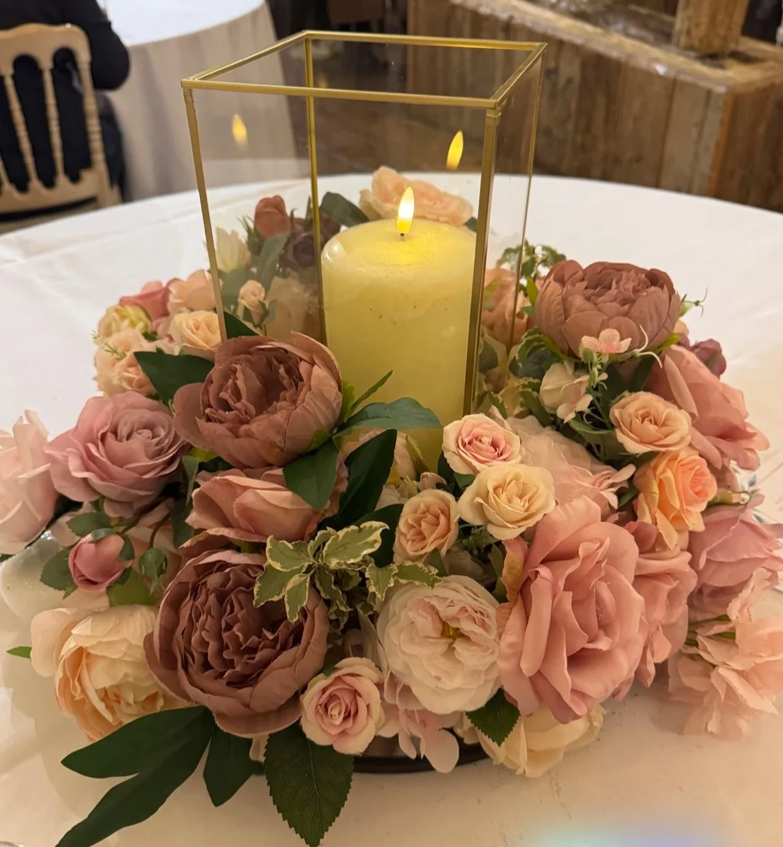 A decorative centerpiece featuring a candle encased in a glass structure surrounded by various flowers in soft colors.