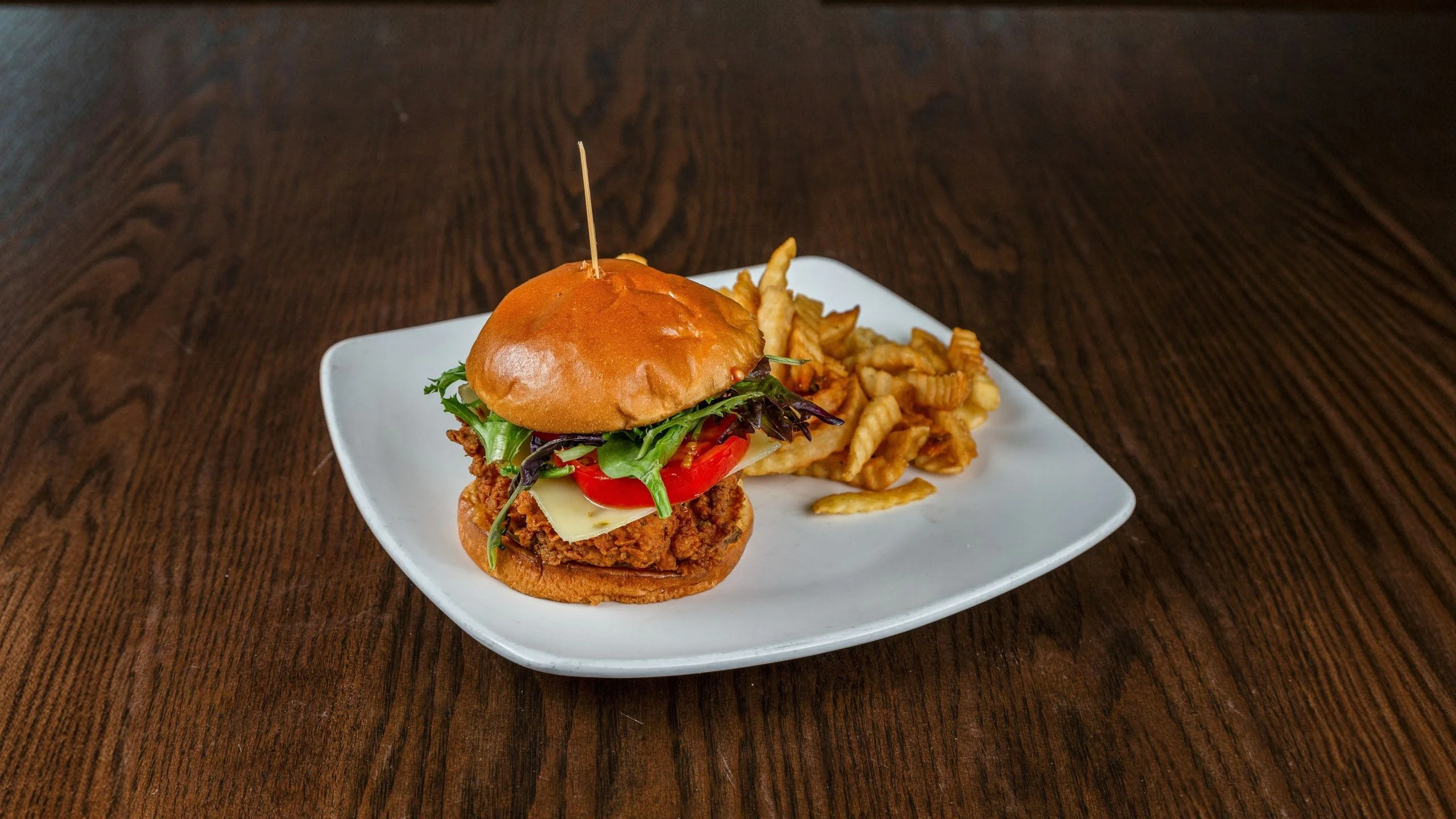 A crispy chicken sandwich topped with lettuce, tomato, and cheese, served with crinkle-cut fries on a white plate.