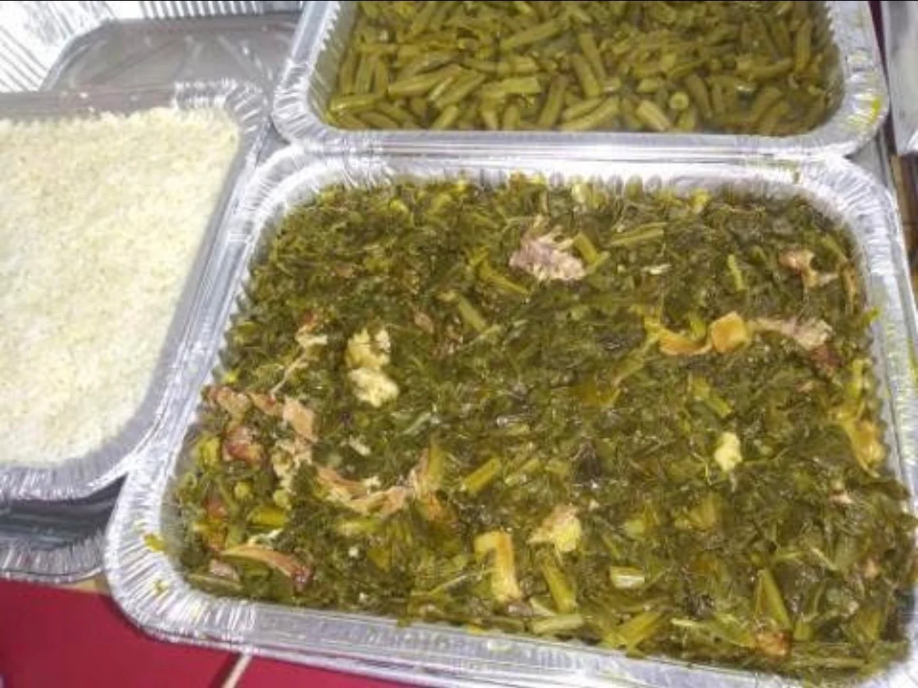 A tray of cooked greens served alongside a tray of white rice. The greens are mixed with pieces of meat and are displayed in a silver container.