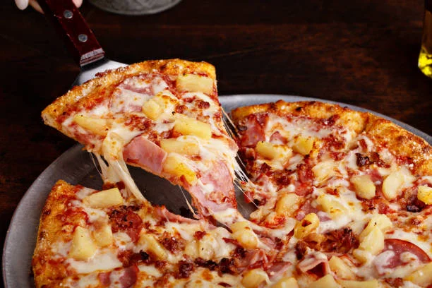 A slice of pizza being lifted from a round pizza with melted cheese, ham, and pineapple toppings.