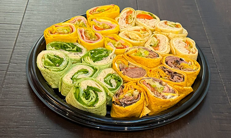 A black dish filled with a creamy salad mixture containing diced ham, diced salami, vegetables, and garnished with seasoning, and surrounded by slices of white bread on a black platter with a decorative lace doily. The platter is placed on a table with a golden tablecloth.