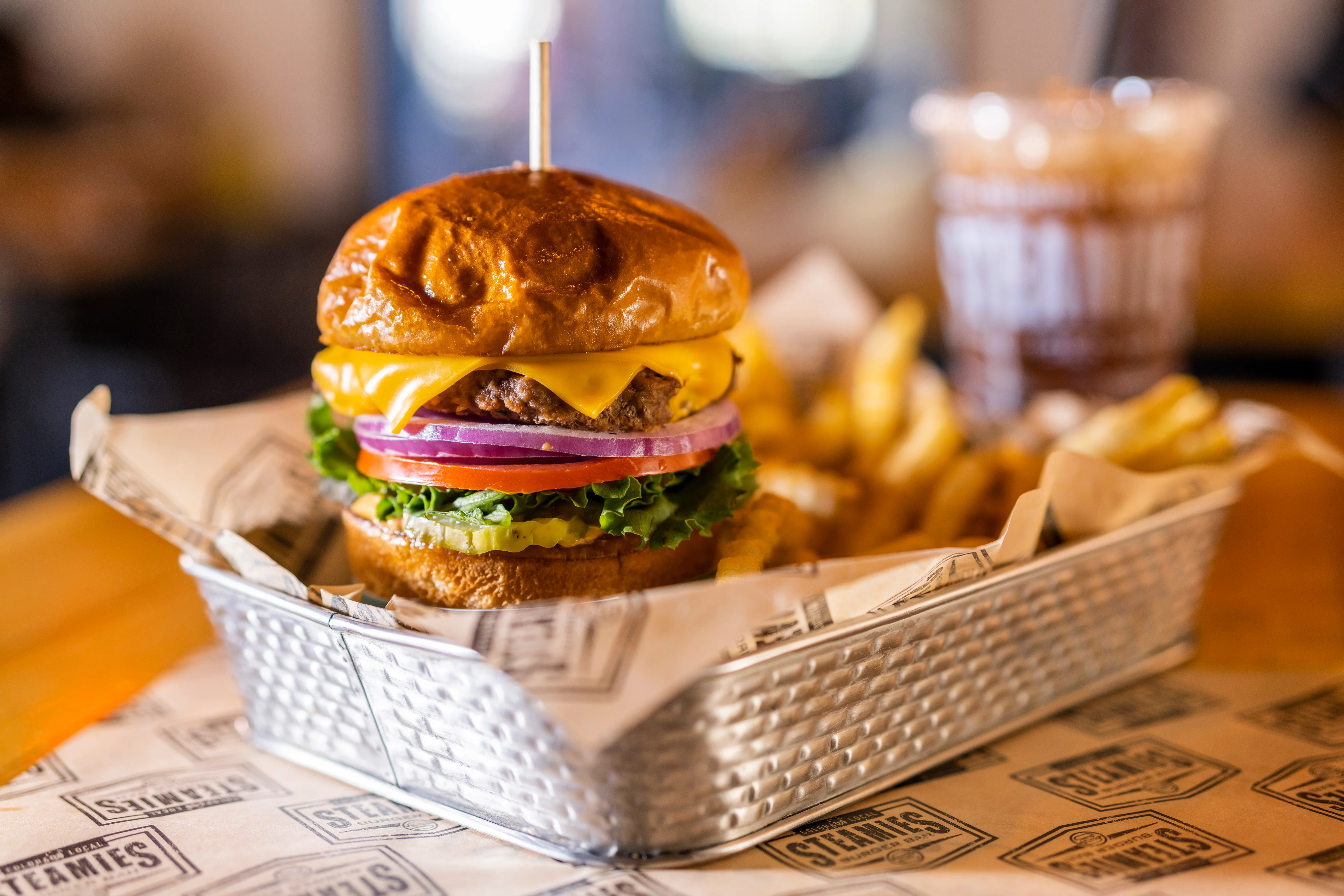 A hearty cheeseburger with layers of lettuce, tomato, onion, and pickles, served with crinkly fries in a metal basket.