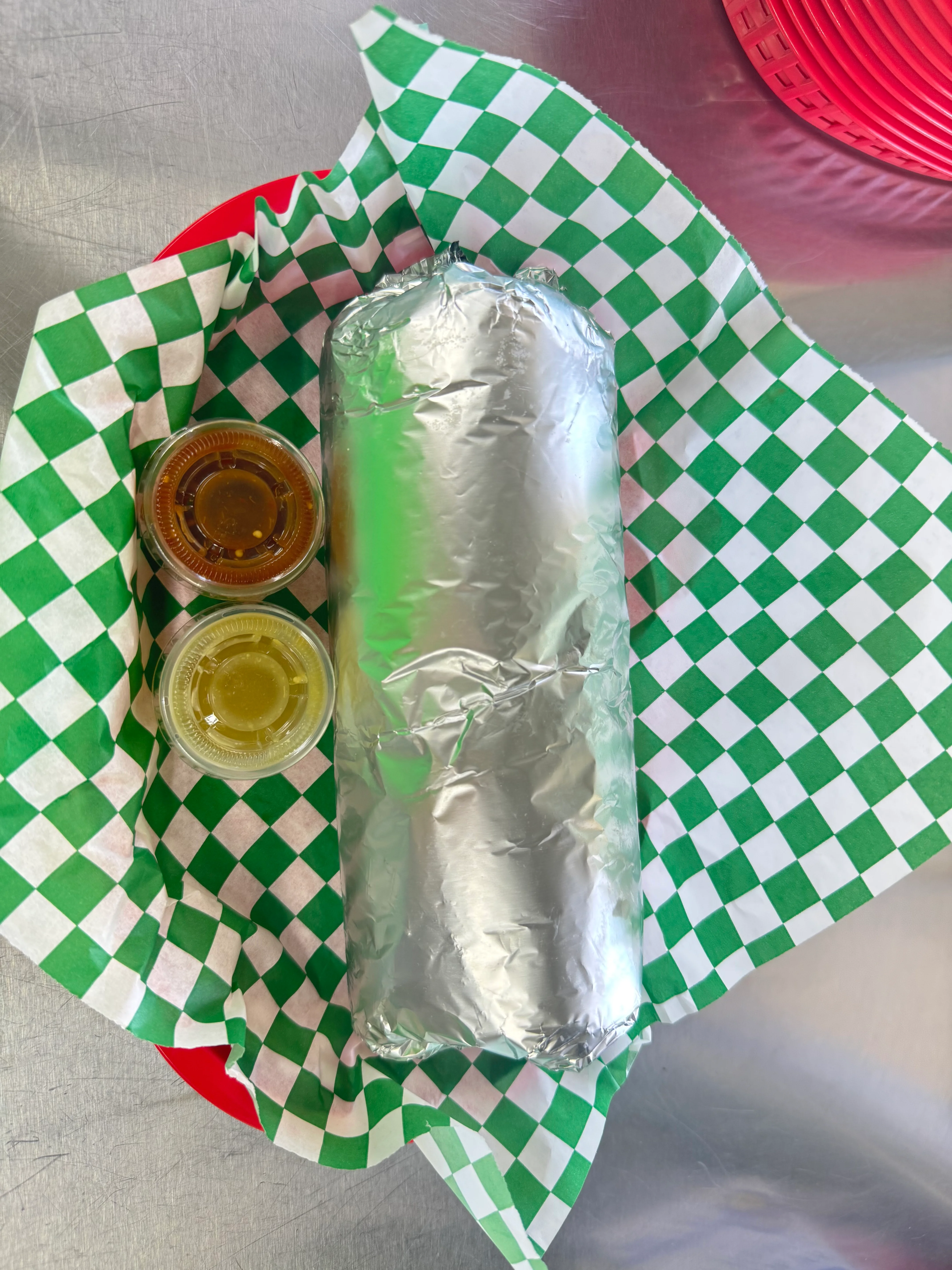 A wrapped burrito placed on a green and white checkered paper, accompanied by two small containers of sauce.