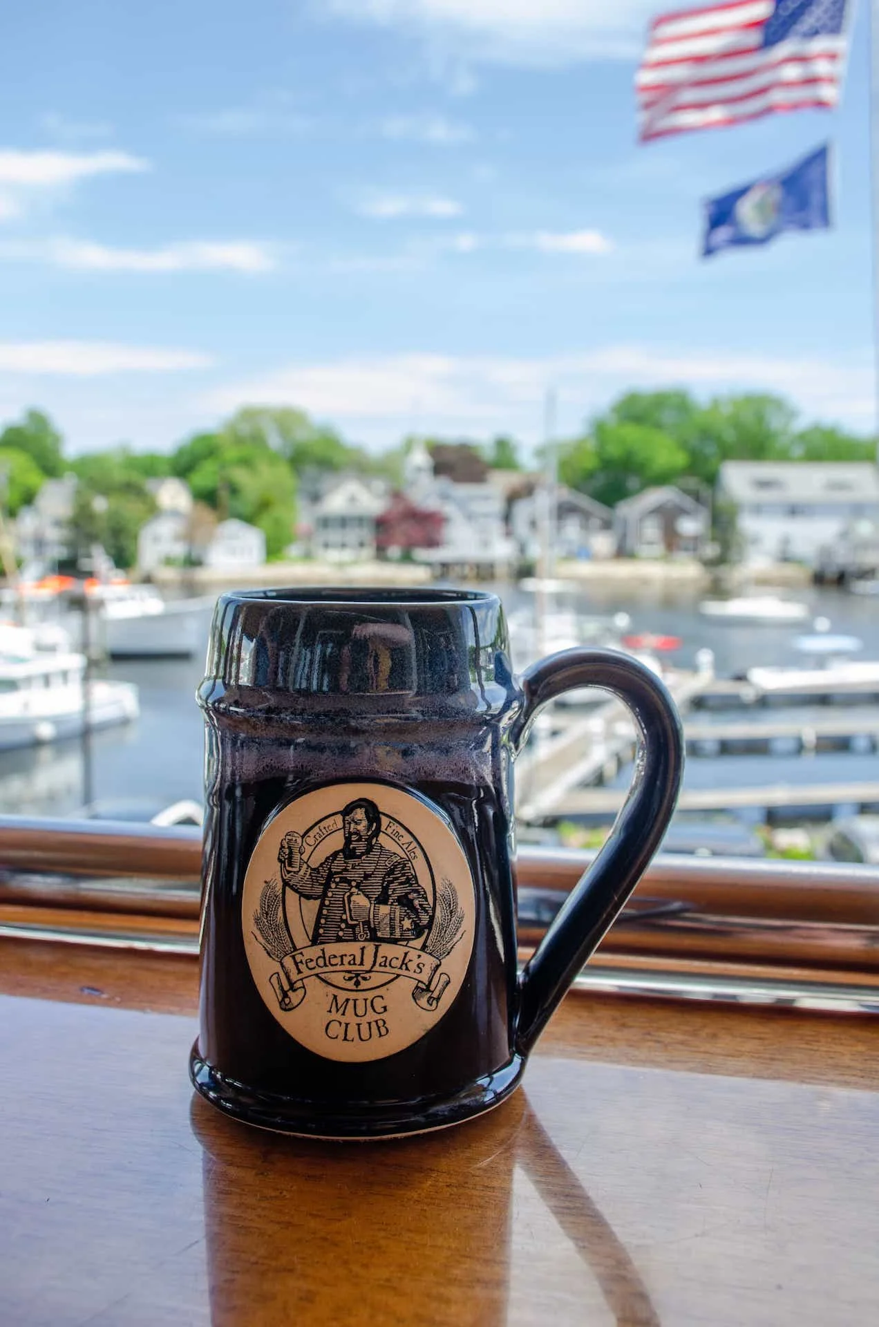 A dark beer mug featuring an illustration and text. The mug is placed on a wooden surface, with a scenic view of a marina and trees in the background.