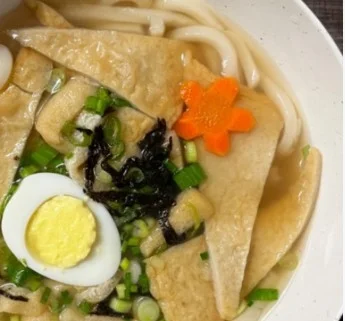 A bowl of noodle soup featuring wide udon noodles, pieces of tofu, green onions, boiled egg, and garnished with a carved carrot flower and seaweed.