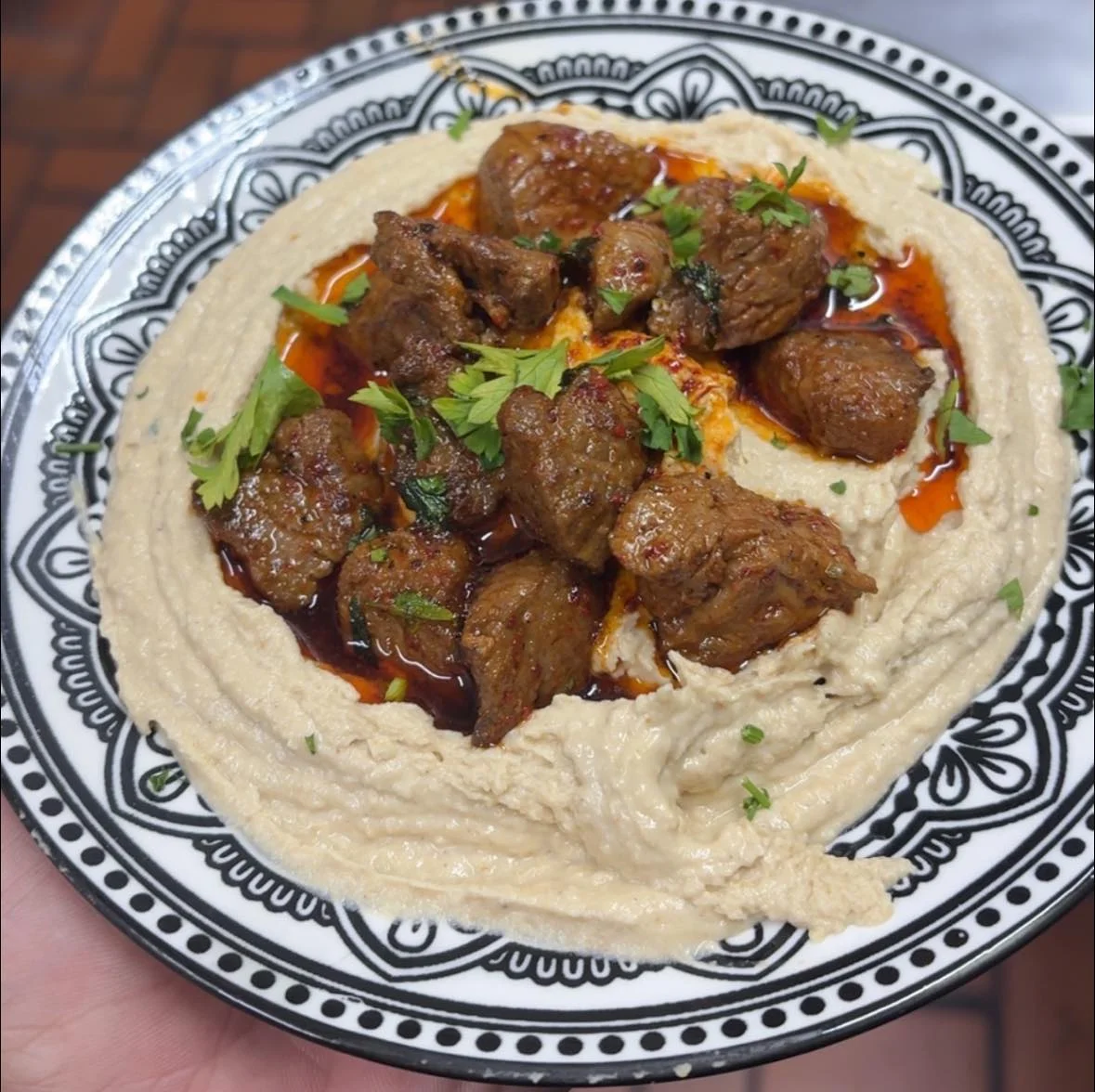 A plate of hummus topped with chunks of seasoned beef, garnished with fresh herbs and a drizzle of oil. The dish is presented with a decorative plate.