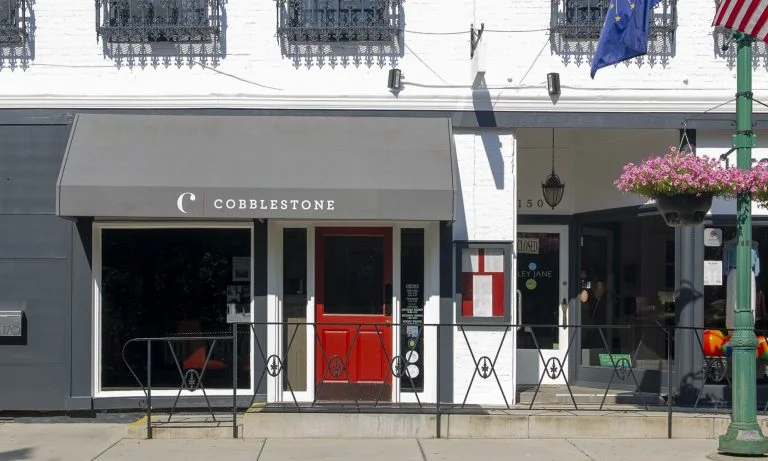 The exterior of a restaurant named Cobblestone, featuring a prominent red door and a covered awning. The façade displays large windows, and decorative elements are present on the building.