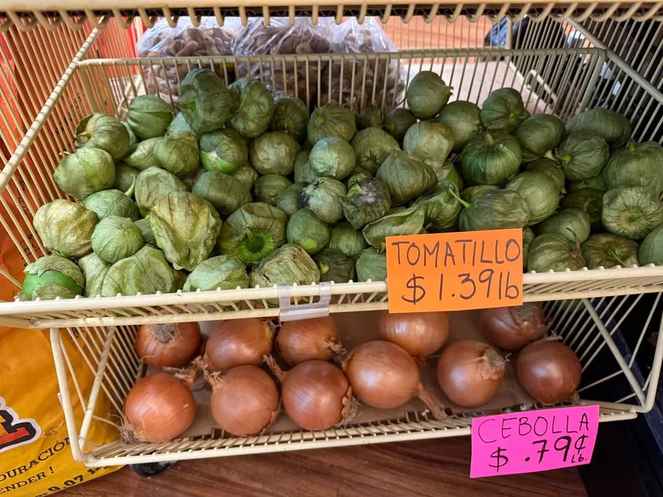 A display of fresh tomatillos and onions in a basket, with price tags indicating their cost per pound.