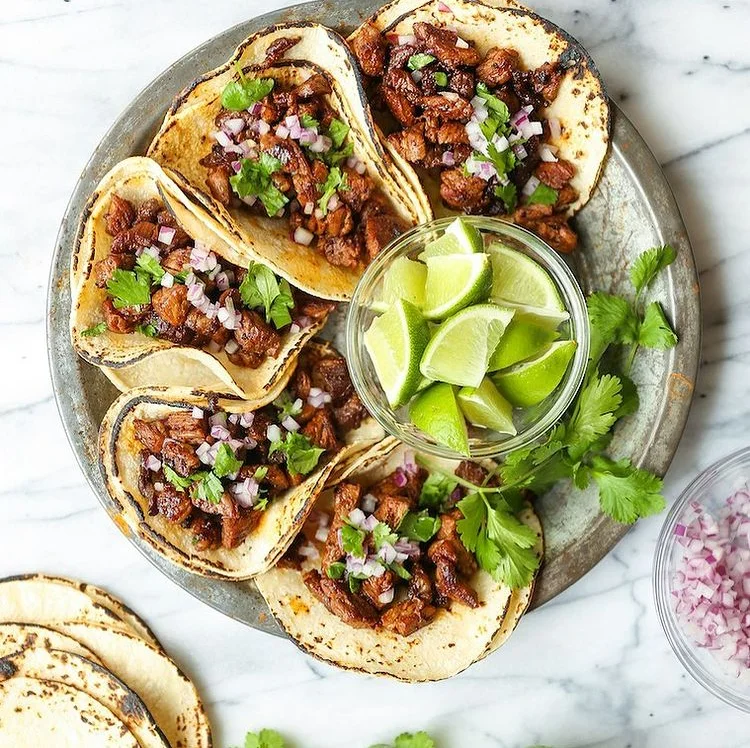 A platter of tacos filled with seasoned meat and topped with onions and cilantro, accompanied by a bowl of lime wedges for garnish.