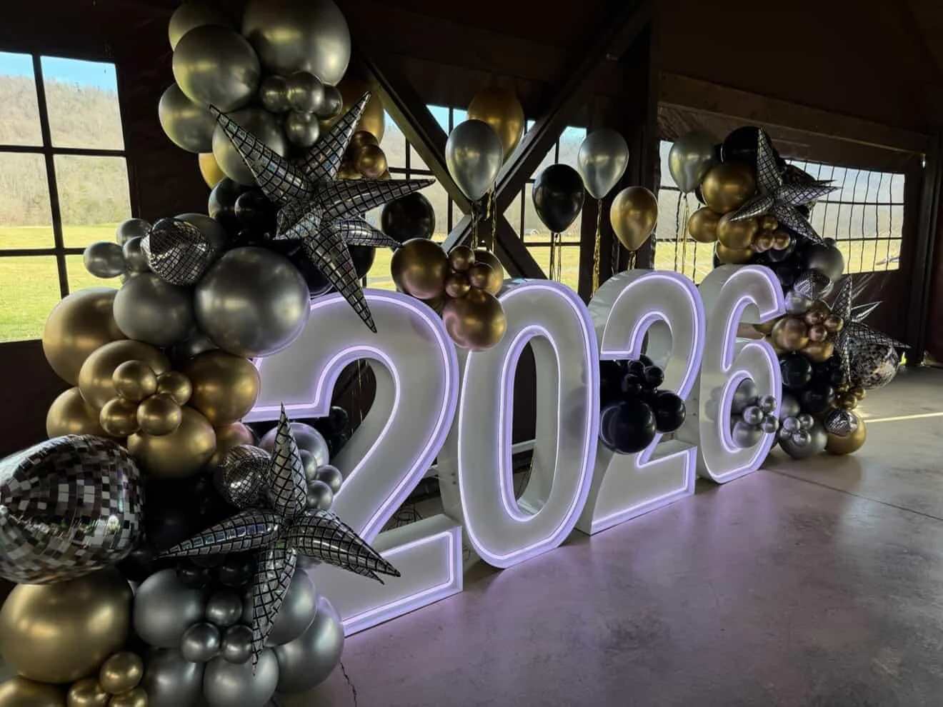 A decorative display featuring large illuminated numbers '2026' surrounded by a cluster of balloons in gold, silver, and black.