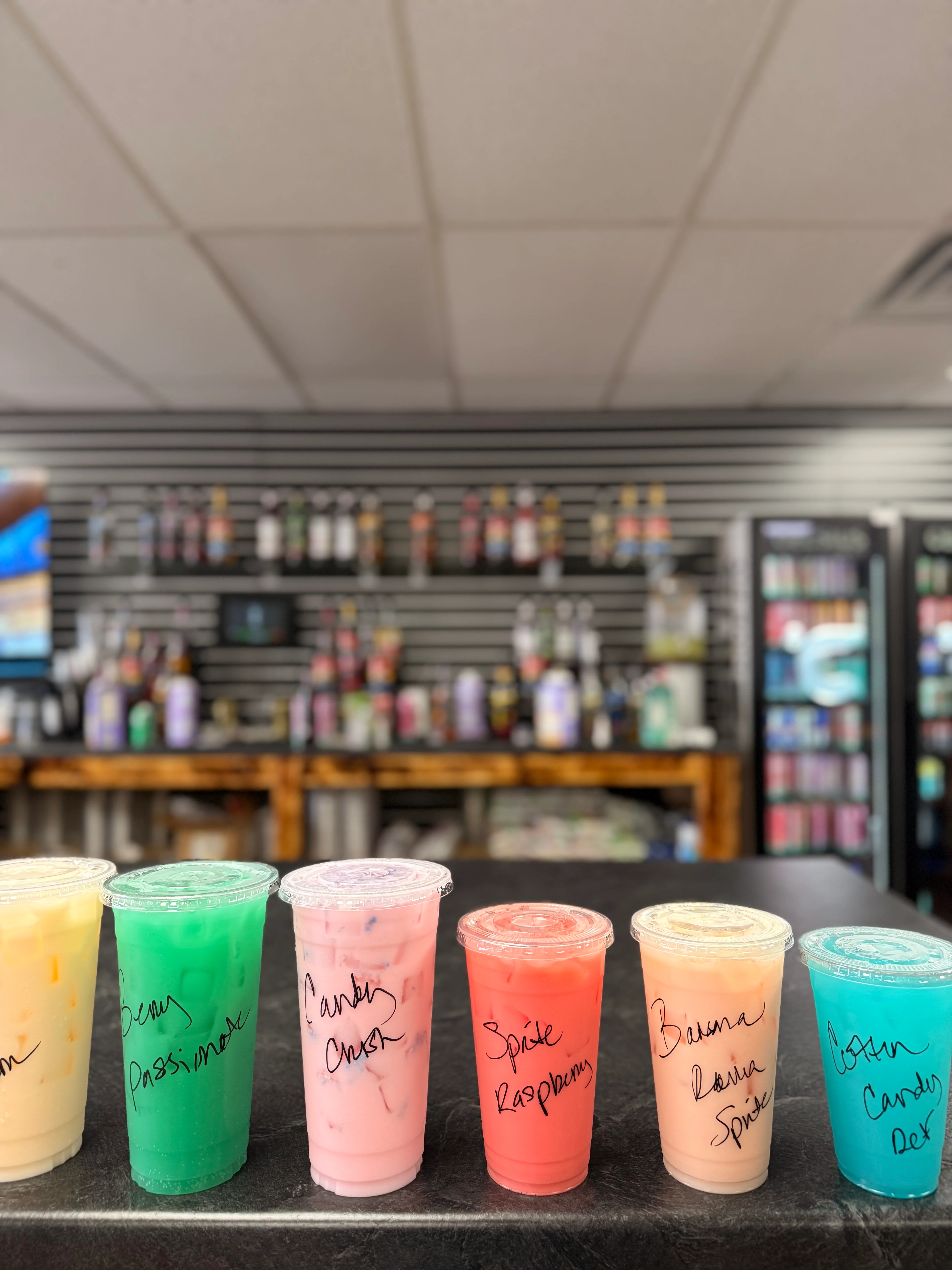 A selection of six drinks in clear cups, each labeled with unique names, displayed on a countertop.