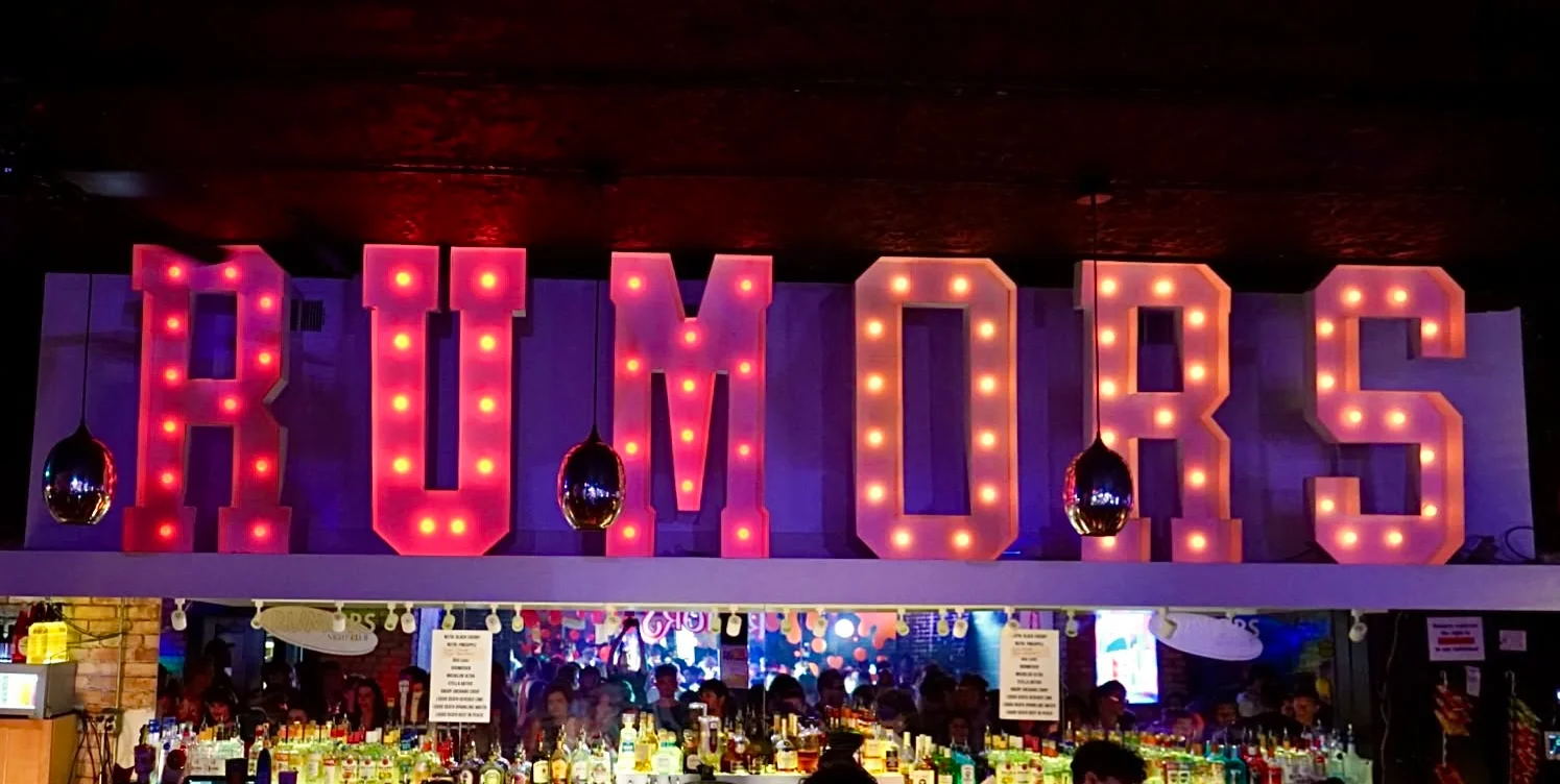 A person making a playful gesture at a bar with drinks in the background.