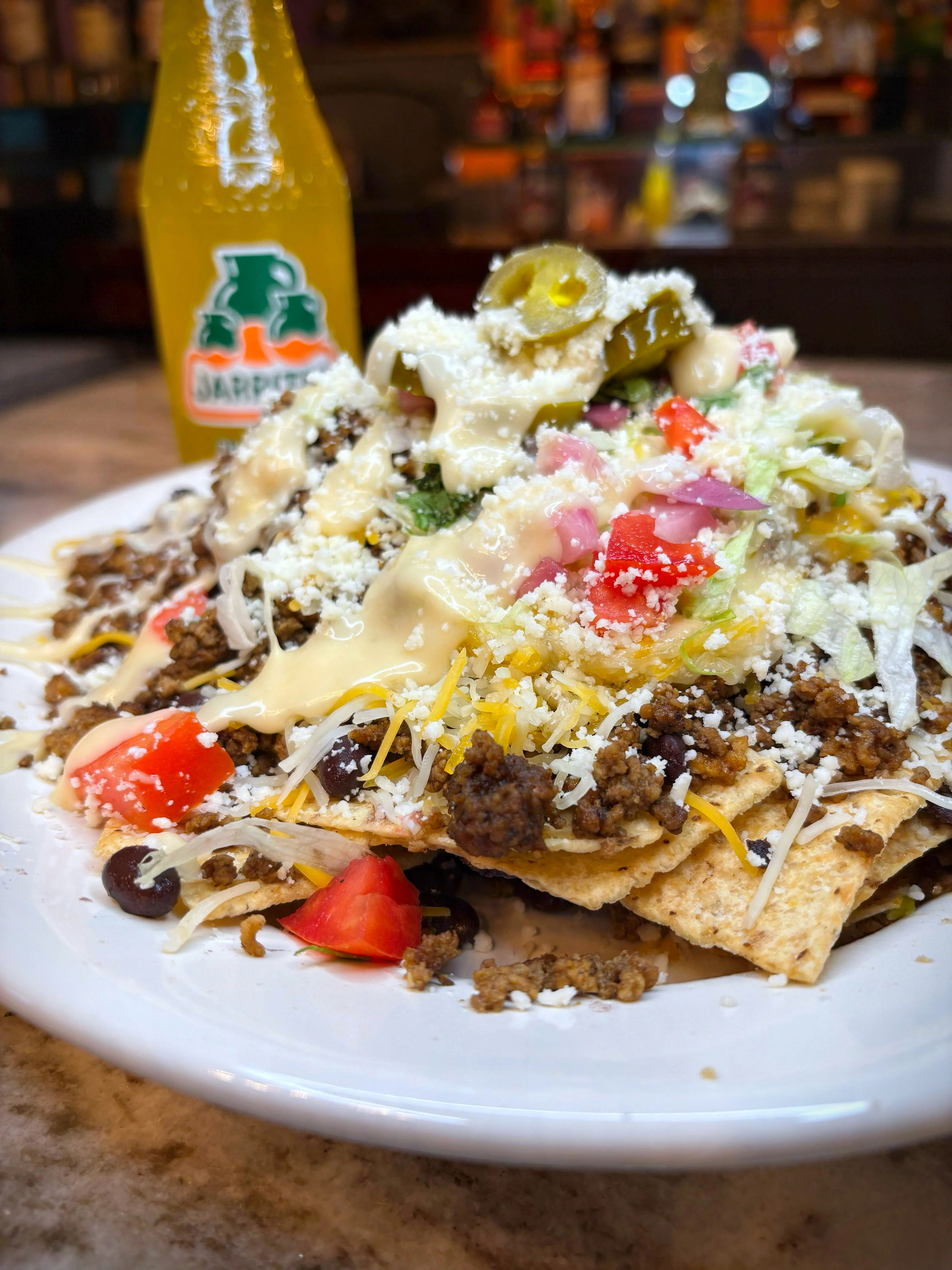 A delicious plate of nachos topped with seasoned meat, cheese, jalapeños, and fresh vegetables. A bottle of soda is in the background.