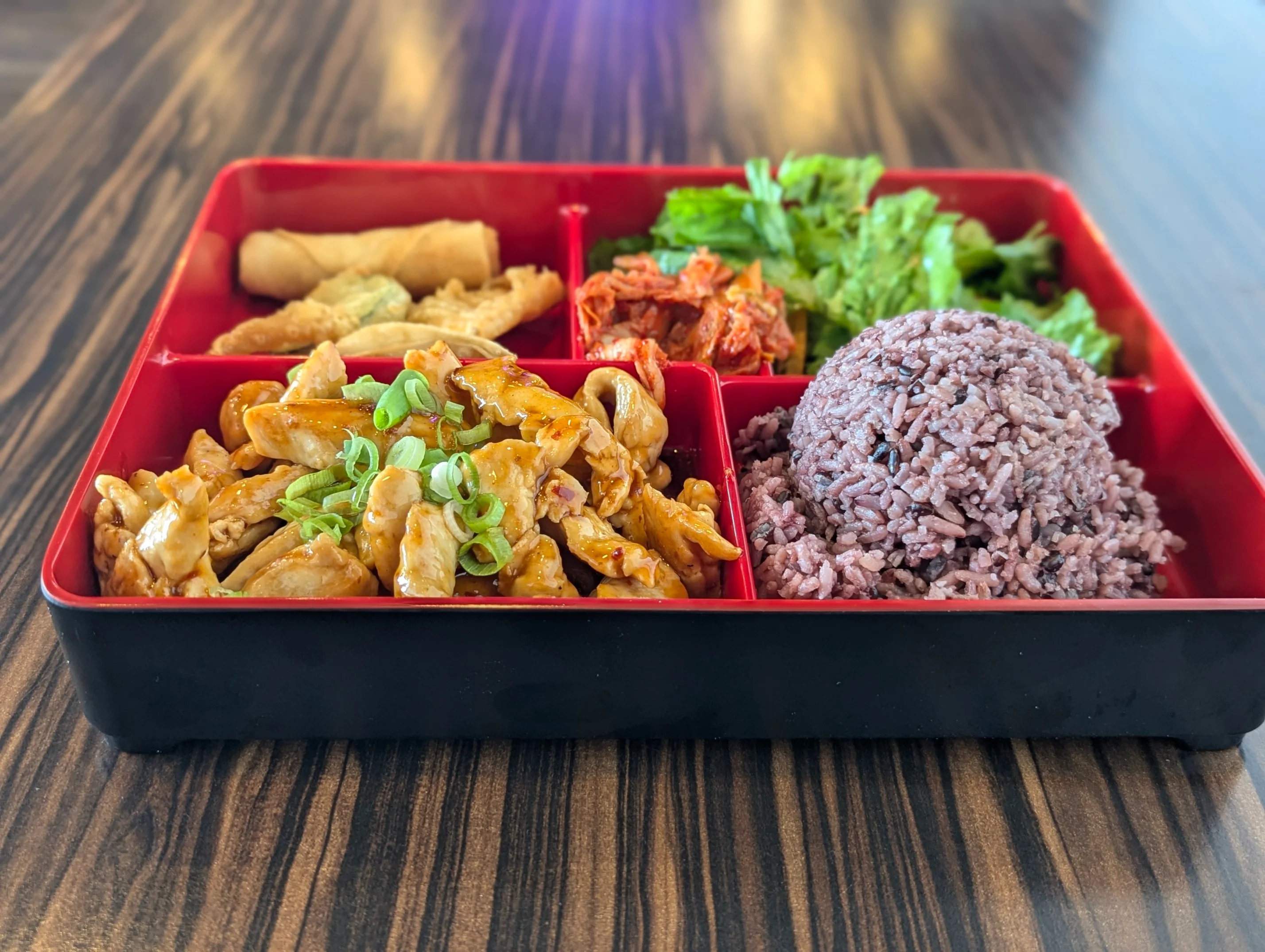 Bento box meal from Bento Kitchen Cravings featuring a portion of purple rice, glazed chicken topped with green onions, kimchi, fresh greens, and assorted crispy appetizers, served on a wooden table for a balanced and visually appealing presentation.