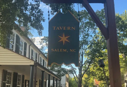 Hanging wooden sign reading “Tavern Salem, NC” along a historic street lined with trees, colonial-style buildings, and parked cars.