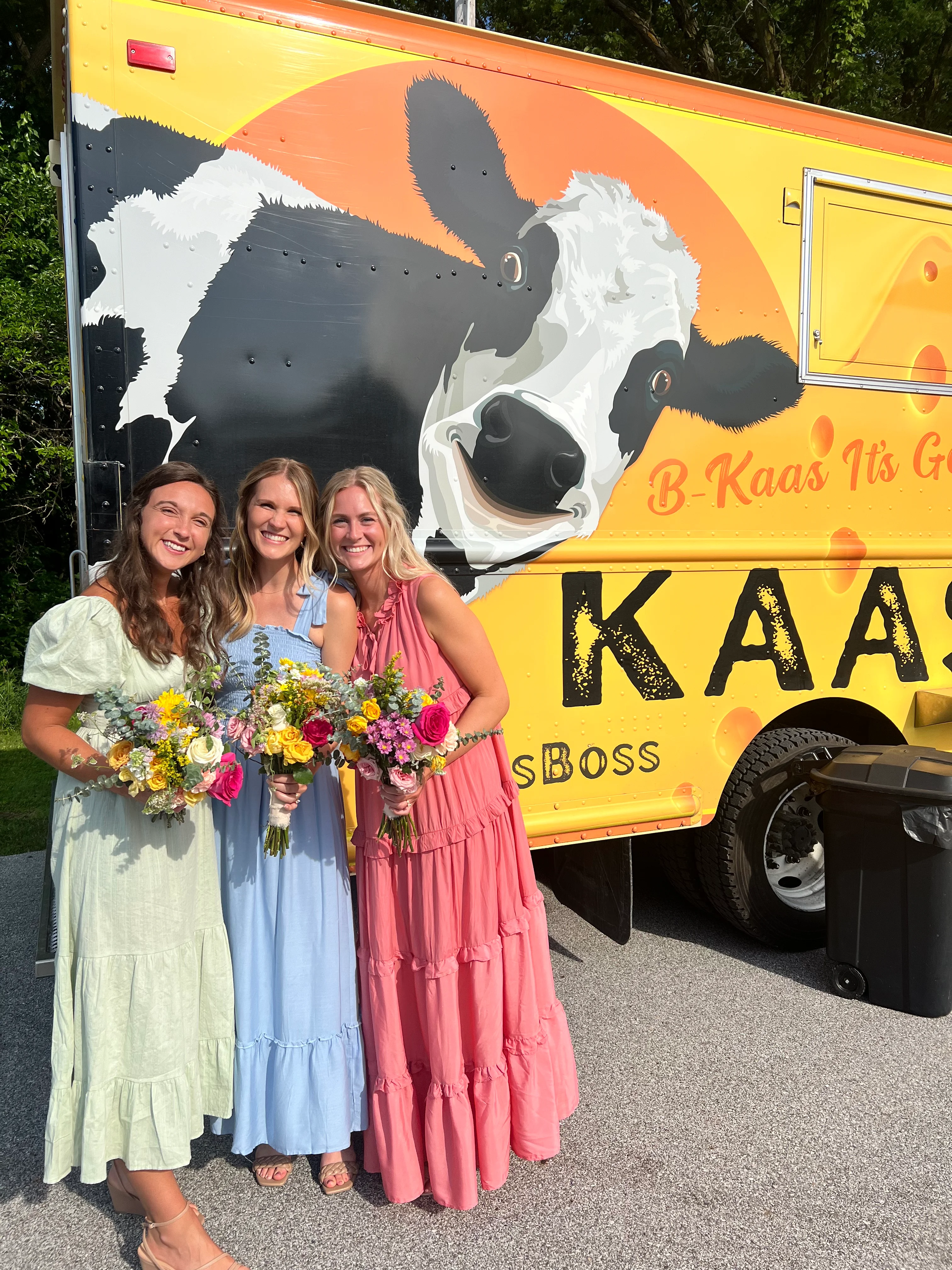Three women in colorful dresses with bouquets by a food truck