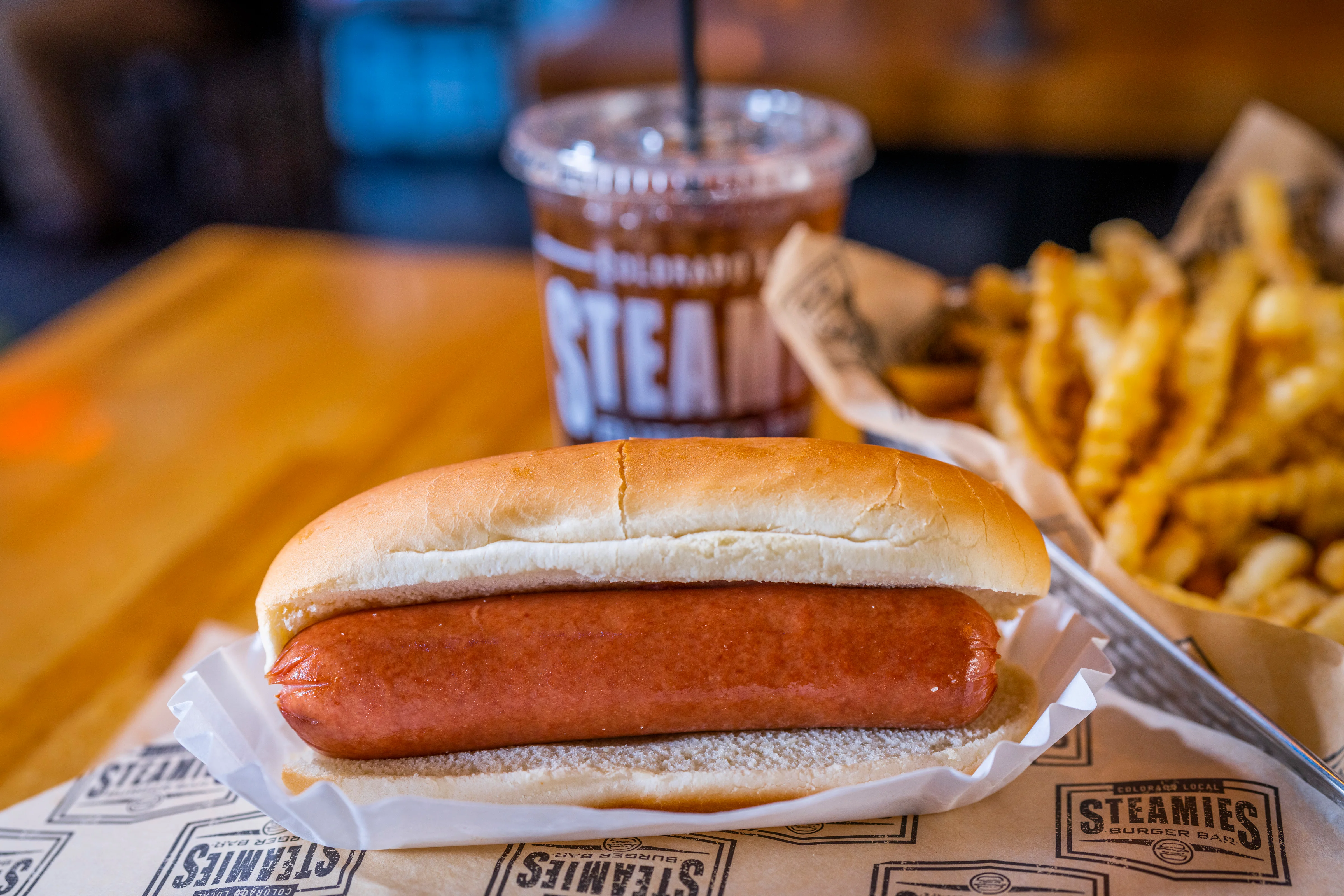 A hot dog in a bun served with a drink and a side of fries in a rustic setting.