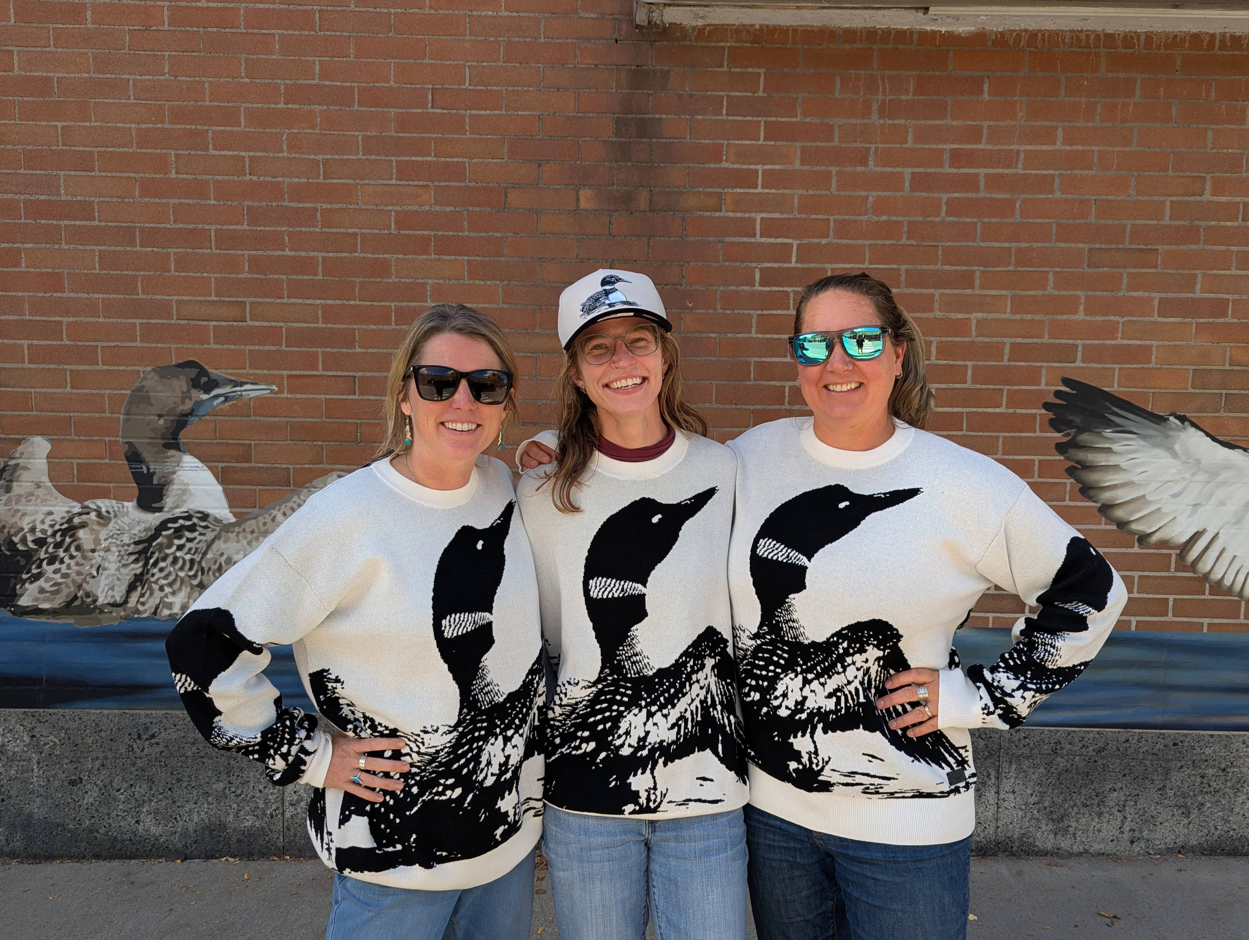 Three friends in matching loons sweaters posing in front of a mural