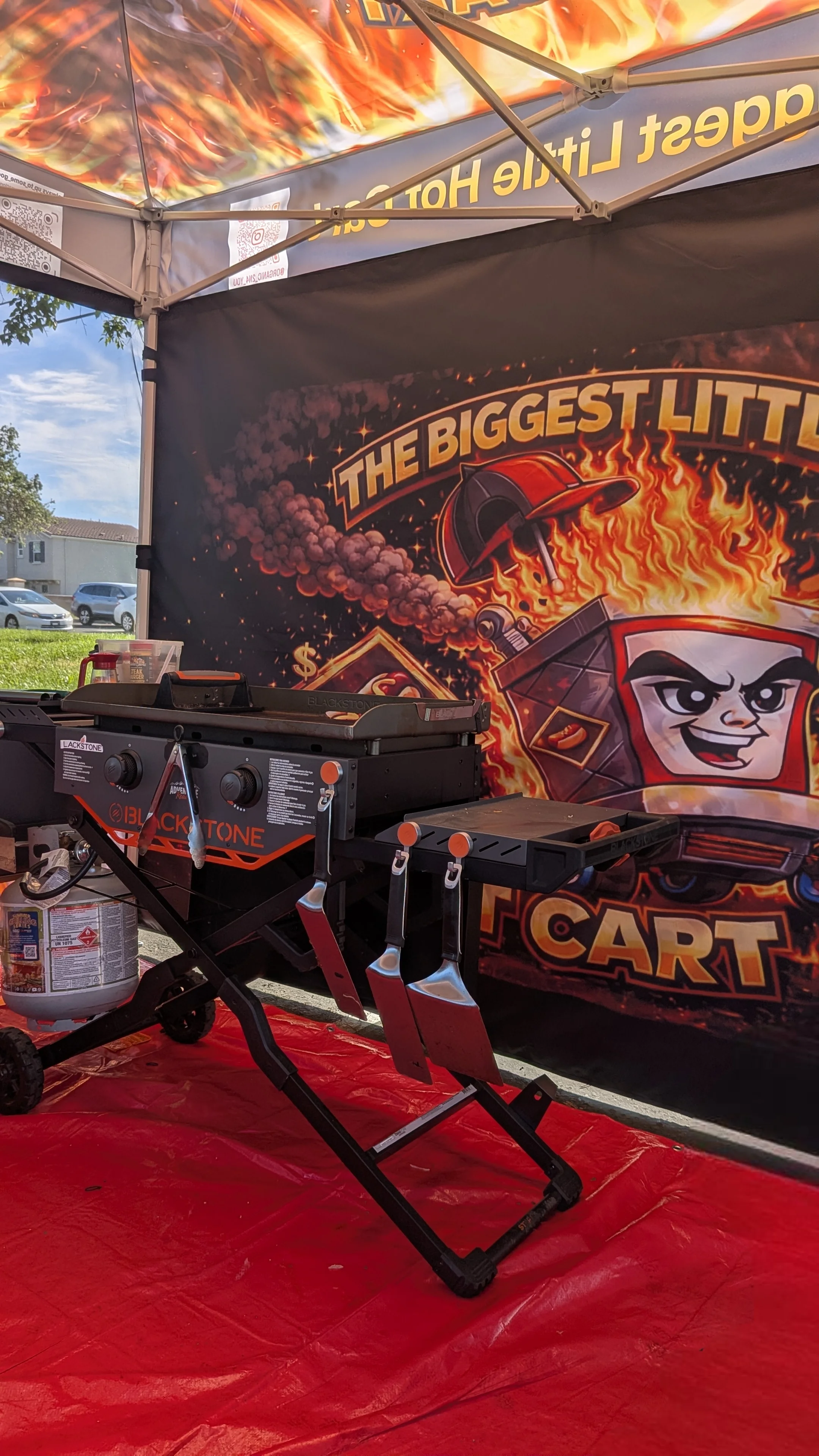 A portable griddle set up under a tent with a bold graphic backdrop featuring flames and a cartoon character. The griddle is equipped with propane and several utensils.