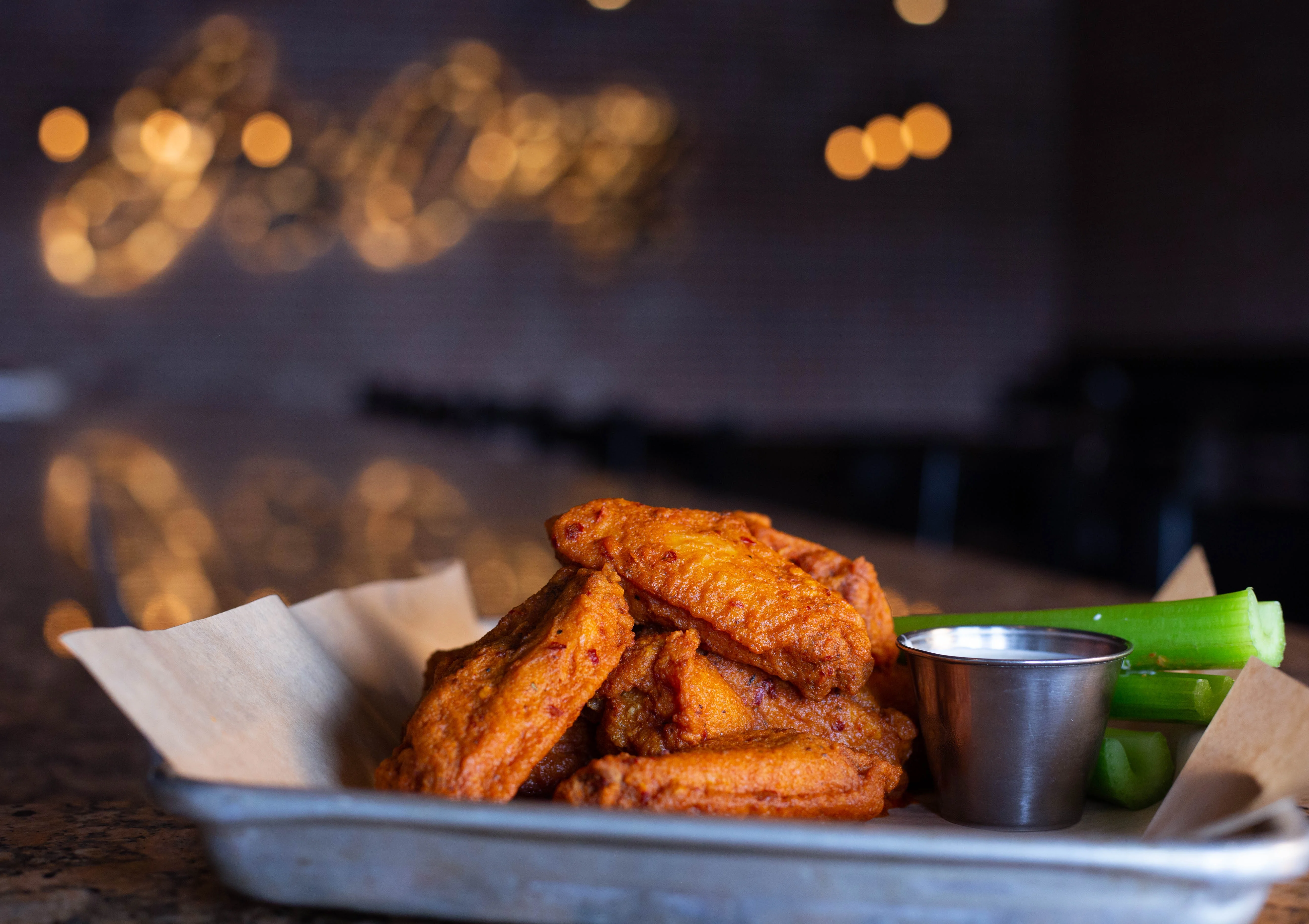 A platter of crispy chicken wings served with celery sticks and a small cup of dipping sauce, set against a blurred restaurant background.