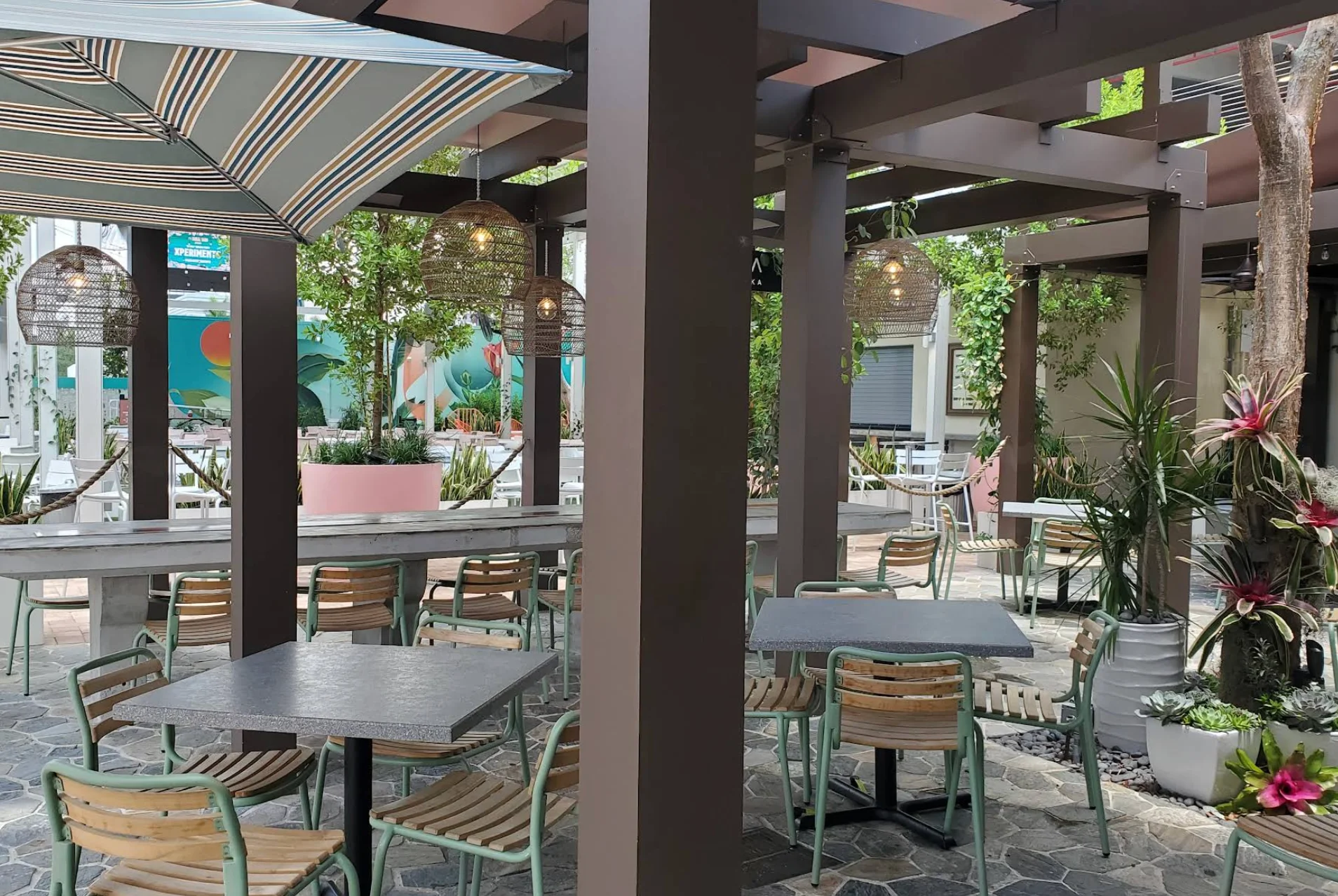 Outdoor patio with modern tables, wooden chairs, and large striped umbrellas. Hanging lanterns add a cozy feel. Surrounding plants provide a lush atmosphere.