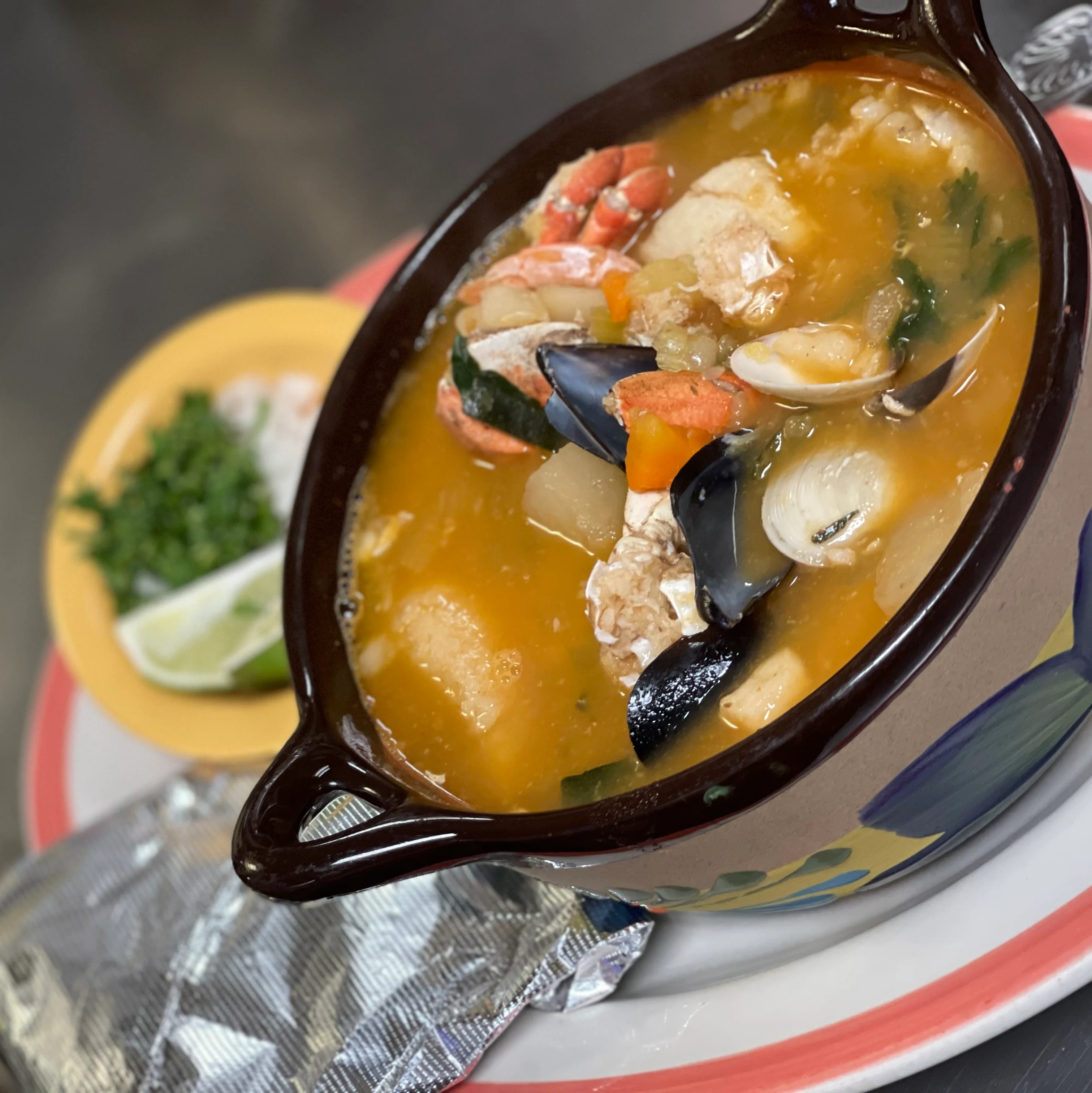 A bowl of seafood soup filled with various seafood, vegetables, and accompanied by a slice of lime and chopped herbs on the side.
