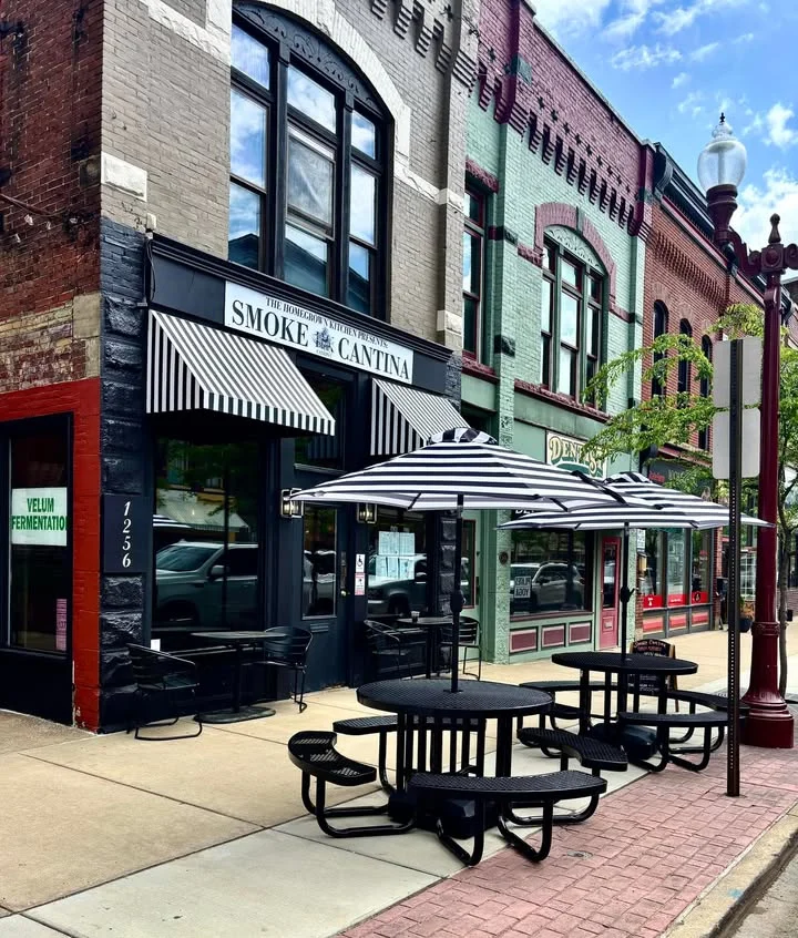 An outdoor view of a restaurant with striped umbrellas over black tables and chairs, set against a backdrop of historic brick buildings.