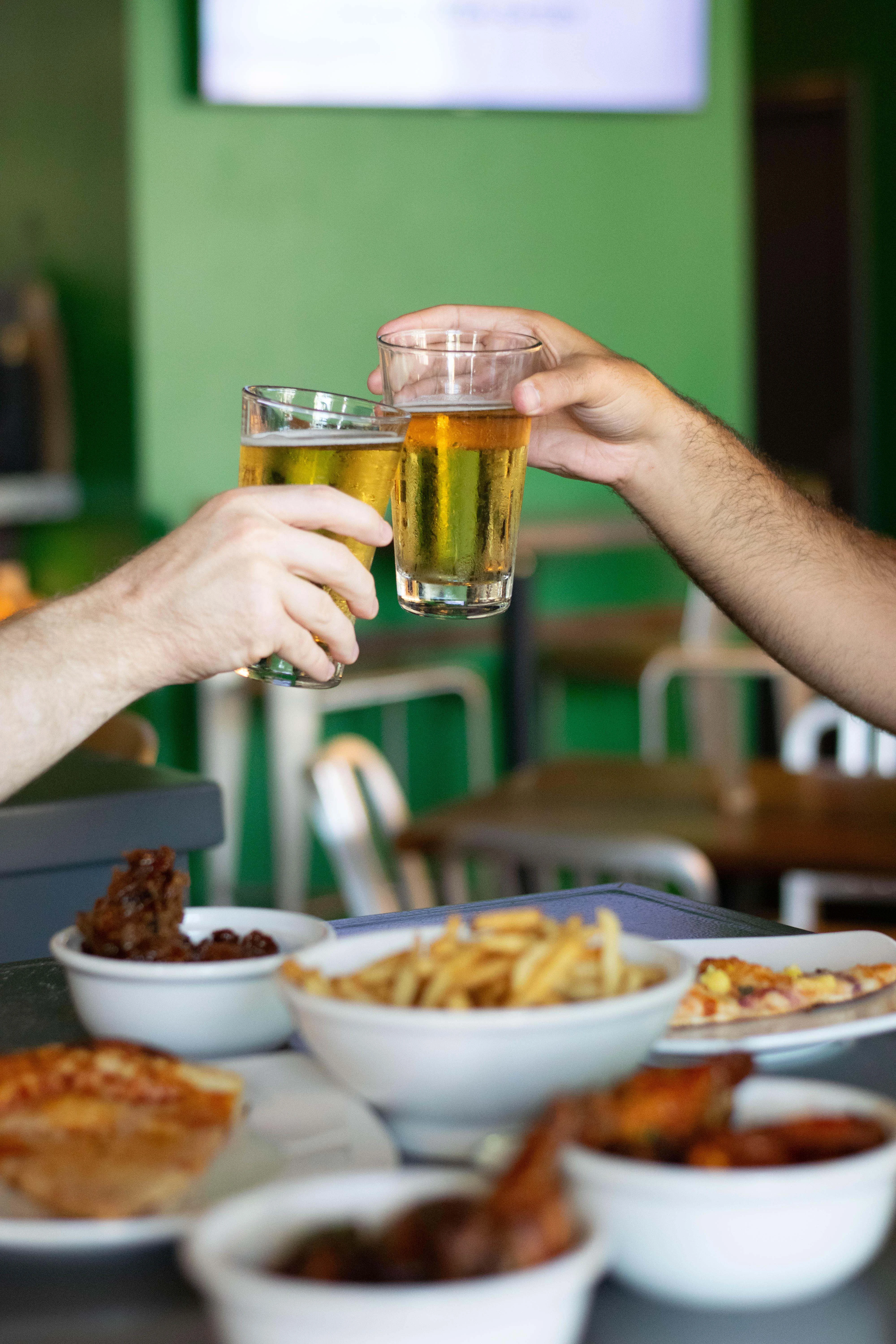 Two glasses of beer are clinking together in celebration over a table filled with appetizers including fries and wings.