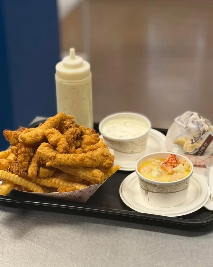 A tray filled with fried chicken tenders, crispy French fries, a cup of ranch dressing, and a bowl of clam chowder.
