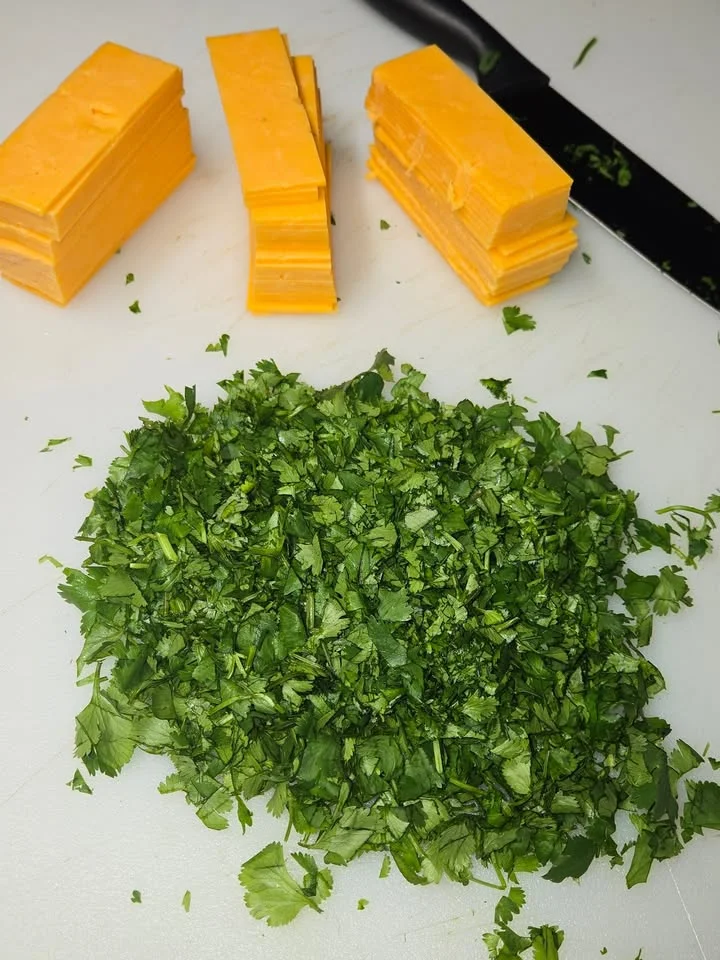 Chopped cilantro neatly arranged on a cutting board, with slices of cheddar cheese in the background.