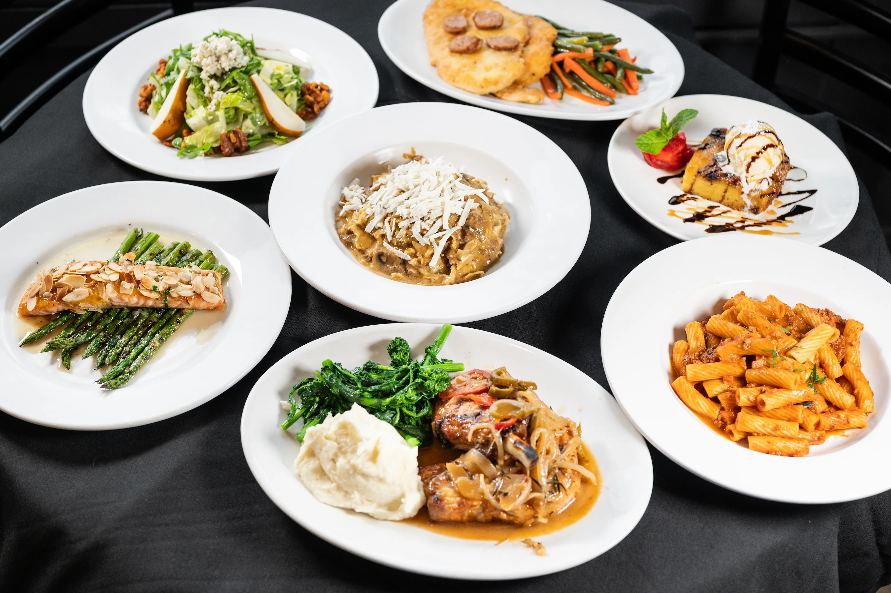 Seven white plates with various dishes, including pasta, fish, chicken, mashed potatoes, vegetables, and a dessert, arranged on a black tablecloth served at Toscana on Main