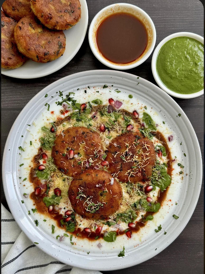 A plate of savory patties served on a bed of yogurt, garnished with pomegranate, fresh herbs, and a drizzle of tamarind and green chutneys.
