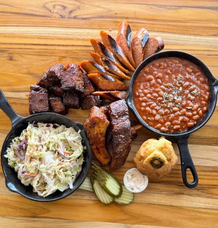 A platter of barbecue meats, sides, and cornbread on a wooden table.